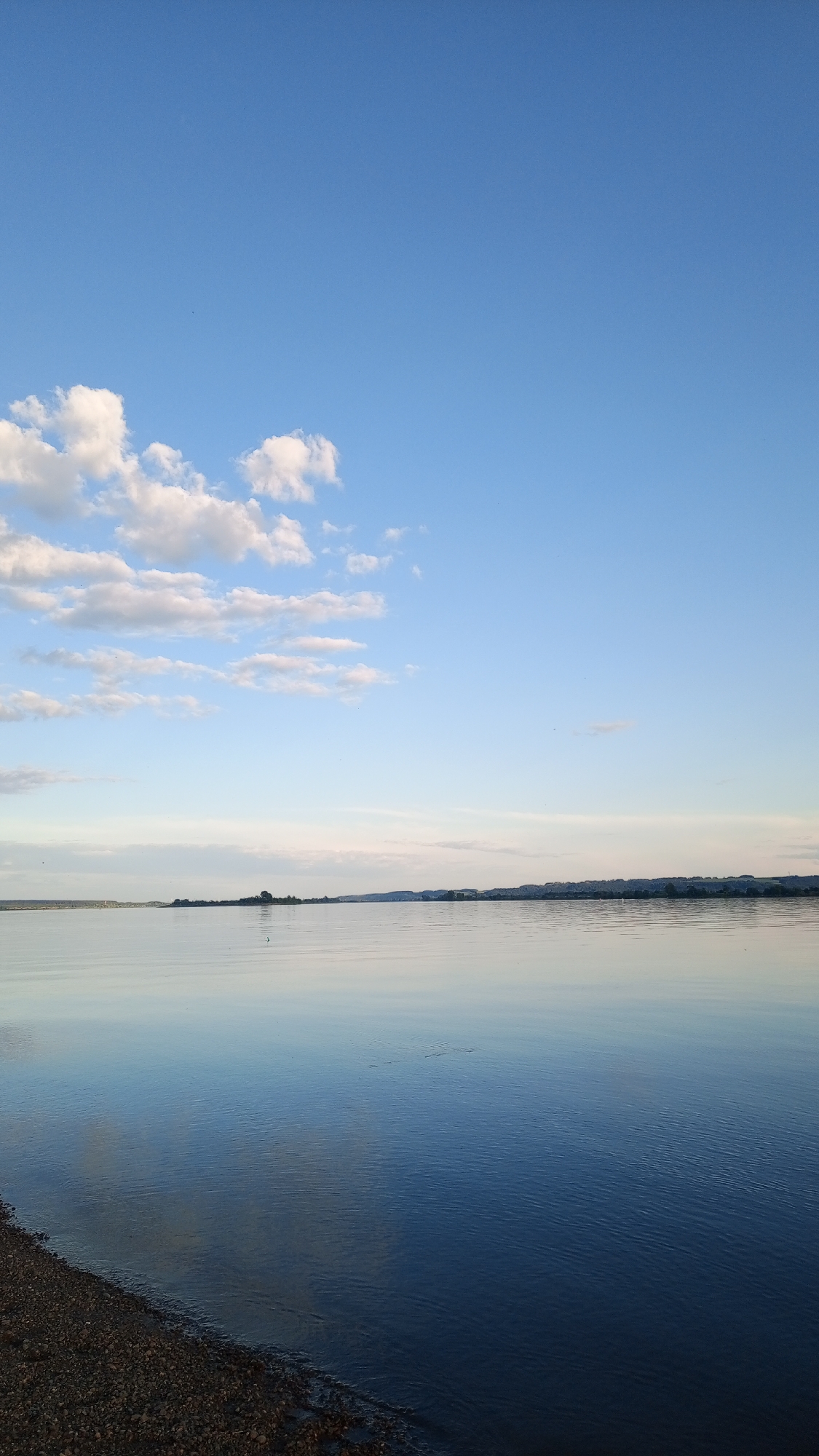 The image displays a landscape view of a wide body of water under a clear blue sky, captured in Шолья, Russia. The upper two-thirds of the frame depict a bright blue sky, which lightens towards the horizon. Scattered white cumulus clouds are visible, primarily in the upper left quadrant. The lower third of the image features the surface of a calm body of water, exhibiting subtle ripples and reflecting the blue tones of the sky. The water appears darker blue in the immediate foreground, transitioning to a lighter, almost grey-blue towards the distant horizon. A low, continuous line of land, characterized by dark green vegetation or structures, is visible across the distant horizon, separating the water from the sky. In the bottom-left corner, a segment of a dark, pebbled shoreline is visible, marking the immediate edge of the water. No individuals, animals, or specific active subjects are discernible within the frame. A faint, dark, thin vertical object is visible on the water's surface in the mid-ground, slightly left of the image center.