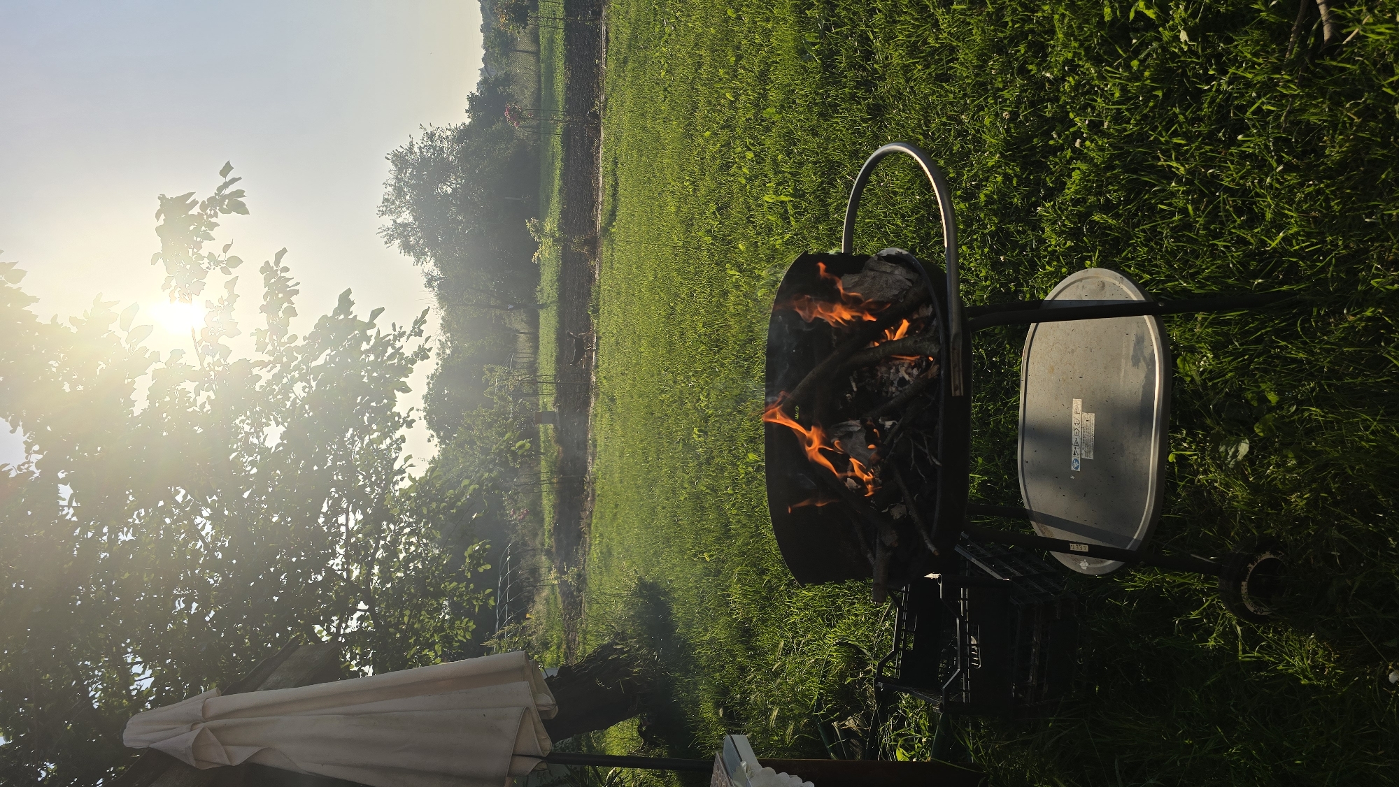 This outdoor image, captured in Rudnik, Bulgaria, depicts a garden or backyard scene under bright sunlight. In the mid-foreground, a dark-colored, portable barbecue grill contains burning wood, producing prominent orange and yellow flames with faint smoke rising. The grill is supported by a stand featuring a lower, grey oval shelf and visible wheels on the left. The ground is covered by green grass, across which long shadows are cast towards the right, indicating a low sun angle. To the immediate left of the grill, two dark plastic crates are visible on the grass. A small portion of a table with a white tablecloth is present in the extreme lower-left corner. In the upper left quadrant, a collapsed, light-colored outdoor umbrella stands against a vertical support, likely a tree trunk. A large tree with green foliage occupies the upper left and center of the frame. The sun, positioned in the upper right quadrant, shines intensely through the tree leaves, creating a bright lens flare and hazy atmospheric effect across the upper portion of the image. Beyond the foreground grass, in the mid-ground, lies an area of cultivated earth showing visible rows of plants and some vertical supports, possibly for climbing plants. A small, dark quadrupedal animal is discernible walking from left to right across this cultivated section. Additional trees and distant vegetation line the background. No human figures are present.