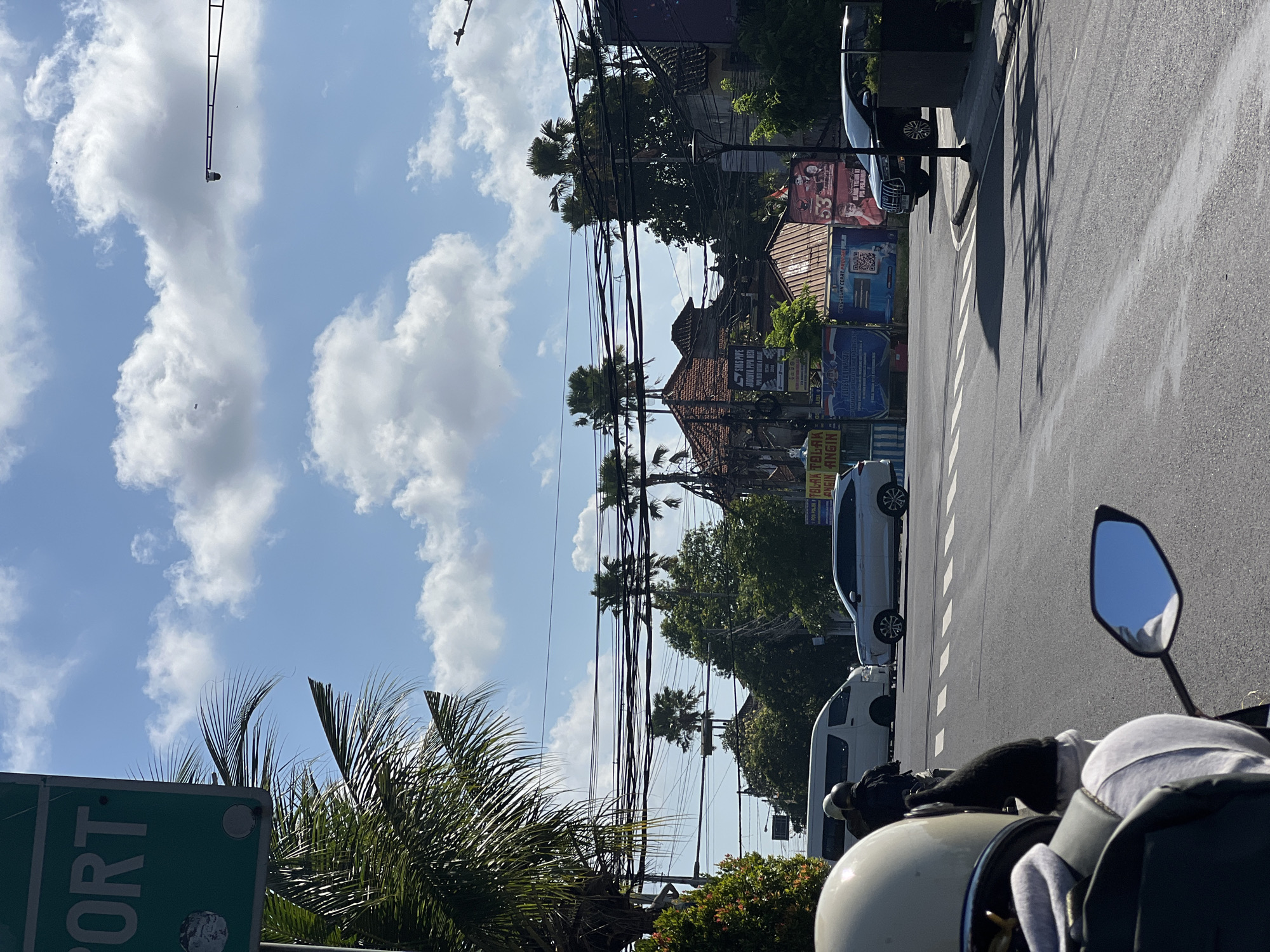 The image shows a street scene in Denpasar, Indonesia, likely near the airport as indicated by a partially visible green sign with the word "PORT" on it. The perspective is from a motorcycle rider, with the back of their helmet and shoulder visible in the foreground. The road is paved with white dashed lines indicating lanes.
Several vehicles are present: a white van, a white SUV, and a black SUV. A person on a motorcycle, wearing a white helmet and dark clothing, is also visible in the distance, in front of the van. A street lamp is visible on the right side of the image.
The background features a mix of tropical vegetation, including palm trees, and buildings with traditional tiled roofs. Overhead, a complex network of electrical wires is strung between poles. The sky is blue with scattered white clouds, suggesting a sunny day. Some advertisements or posters are visible on the buildings in the background.