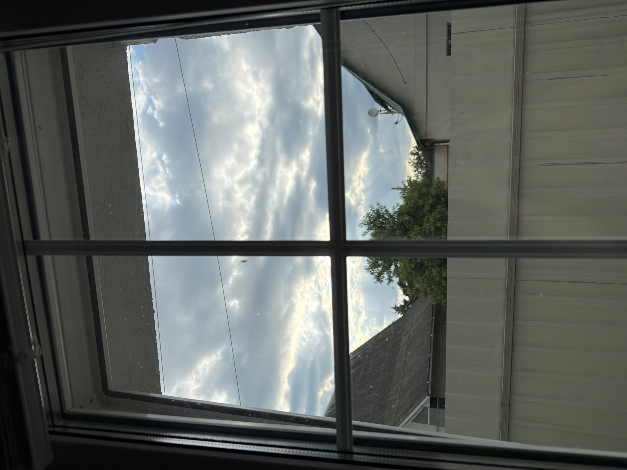 A view through a window shows a cloudy sky with shafts of light breaking through. Below the sky, a grey metal fence runs horizontally across the frame, behind which are rooftops of buildings and trees. One building on the left has a dark grey, corrugated roof. On the right, a light-colored building with a green roof is visible, featuring a satellite dish. The scene appears to be an outdoor area in Bila Tserkva, Ukraine, likely residential. The image is taken from an interior perspective looking outwards through a window with white vertical and horizontal dividers.
