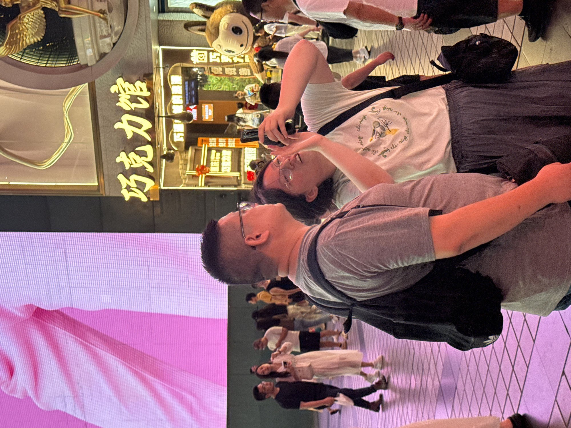 This is a medium shot of a group of people on a city street at night, with a prominent pink digital billboard in the background. The focus is on two individuals in the foreground, a man and a woman. The man, on the left, is wearing a gray t-shirt and a backpack. The woman, in the center, is wearing a white t-shirt with a graphic print and a gray skirt, and is holding a smartphone up, appearing to take a photo. Behind them, a street scene unfolds with other pedestrians, some carrying bags, and shops with illuminated signs, including one with Chinese characters that read "巧克力馆" (Chocolate Museum). The lighting suggests an urban environment with artificial light sources, including streetlights and shop displays. The ground is paved with square tiles. The overall atmosphere is one of a busy commercial area at night.