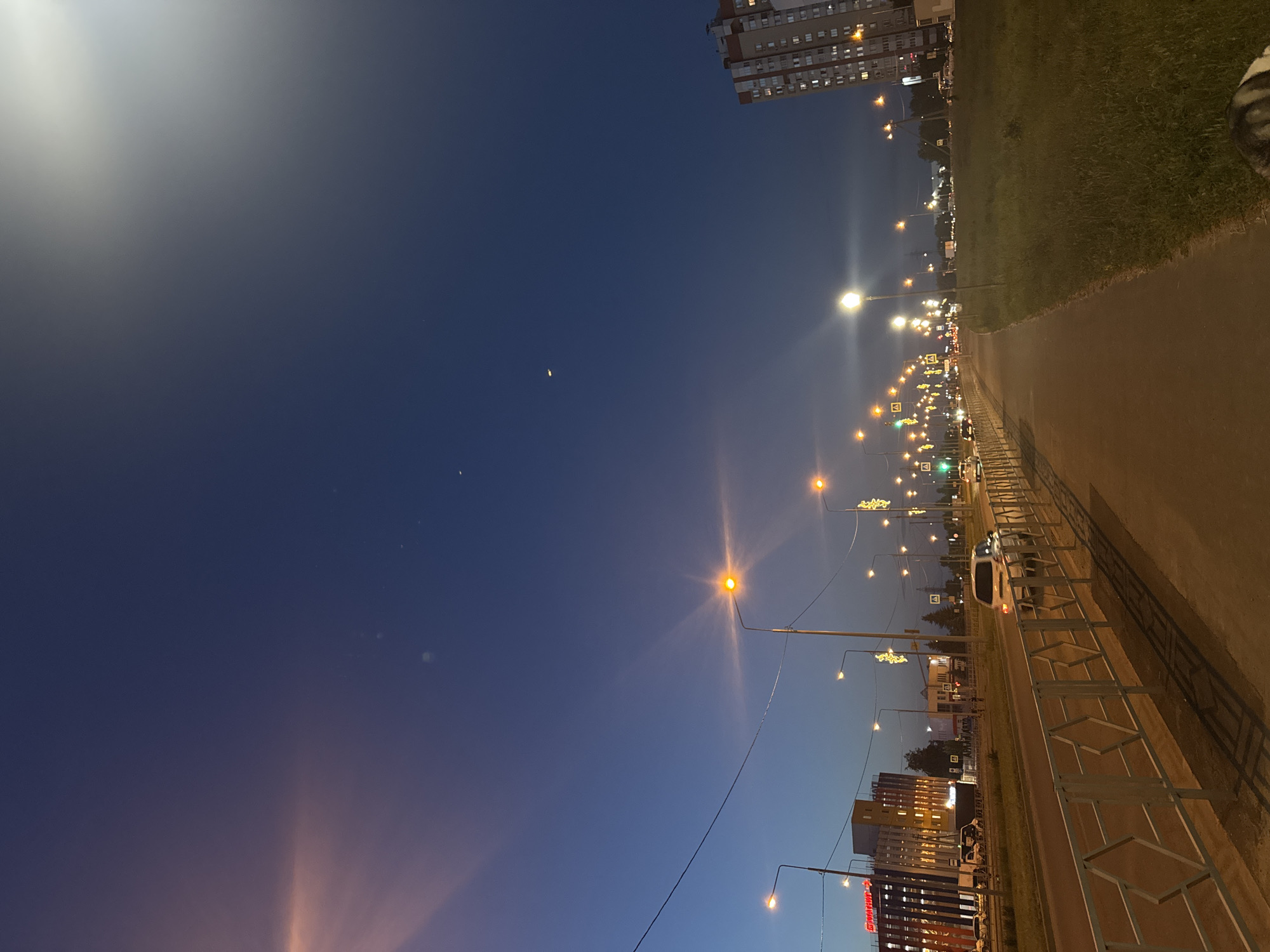 This image depicts an urban street scene at dusk or early night in the city of Unknown, Unknown. The sky transitions from a lighter blue-purple on the left horizon to a deep, dark blue overhead and on the right, suggesting the sun has recently set. Two faint, distant luminous objects are visible in the upper central-right sky; one is crescent-shaped, and the other is a small, bright dot above it.
The scene is illuminated by numerous streetlights emitting warm yellow-orange light, creating prominent lens flares. On the left side of the frame, a multi-lane asphalt road extends into the distance. A white metal barrier with a repetitive hexagonal or diamond-shaped pattern runs alongside a paved sidewalk in the foreground. Beyond the sidewalk and barrier, a white SUV is visible on the road in the mid-ground, heading away from the viewer. Further down the road on the left, a large modern building with a dark lower facade and a yellow/orange upper section features multiple illuminated windows and a prominent red sign with Cyrillic script. Several other vehicles, including a white van, are discernible on the road.
On the right side of the road, a wide, unkempt grassy verge separates the sidewalk from a row of distant trees. Further down the road, additional streetlights are adorned with decorative yellow-gold light strings. In the far background on the right, a tall, multi-story residential building with reddish-brown and white facades has numerous illuminated windows. Traffic lights are visible in the distance, showing green. No individuals are visible within the frame.