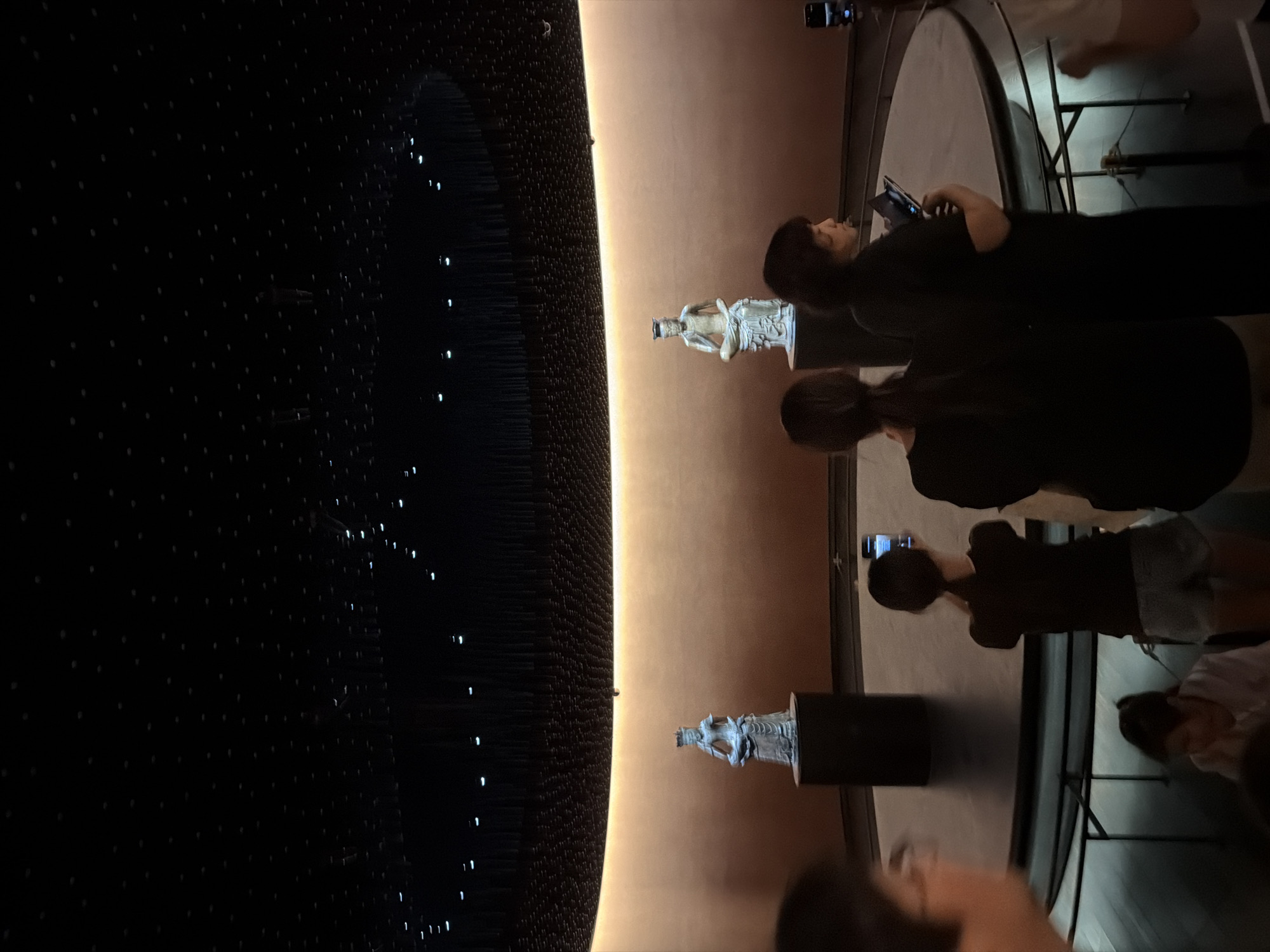 The image depicts the exterior of a circular, dimly lit exhibition space, likely part of a museum or gallery, in Seoul, South Korea. The ceiling is black and adorned with countless small, white lights, resembling stars, creating a celestial effect. A band of warm, golden light emanates from behind a curved wall that forms the backdrop of the exhibition area. Two large, white statues, appearing to be ancient figures, stand on dark cylindrical pedestals in the center of the circular display. One statue is on the left, and the other is on the right. Several people are visible in the foreground, some looking towards the statues and one woman is taking a photo with her phone. The overall ambiance is solemn and contemplative, suggesting a cultural exhibition.