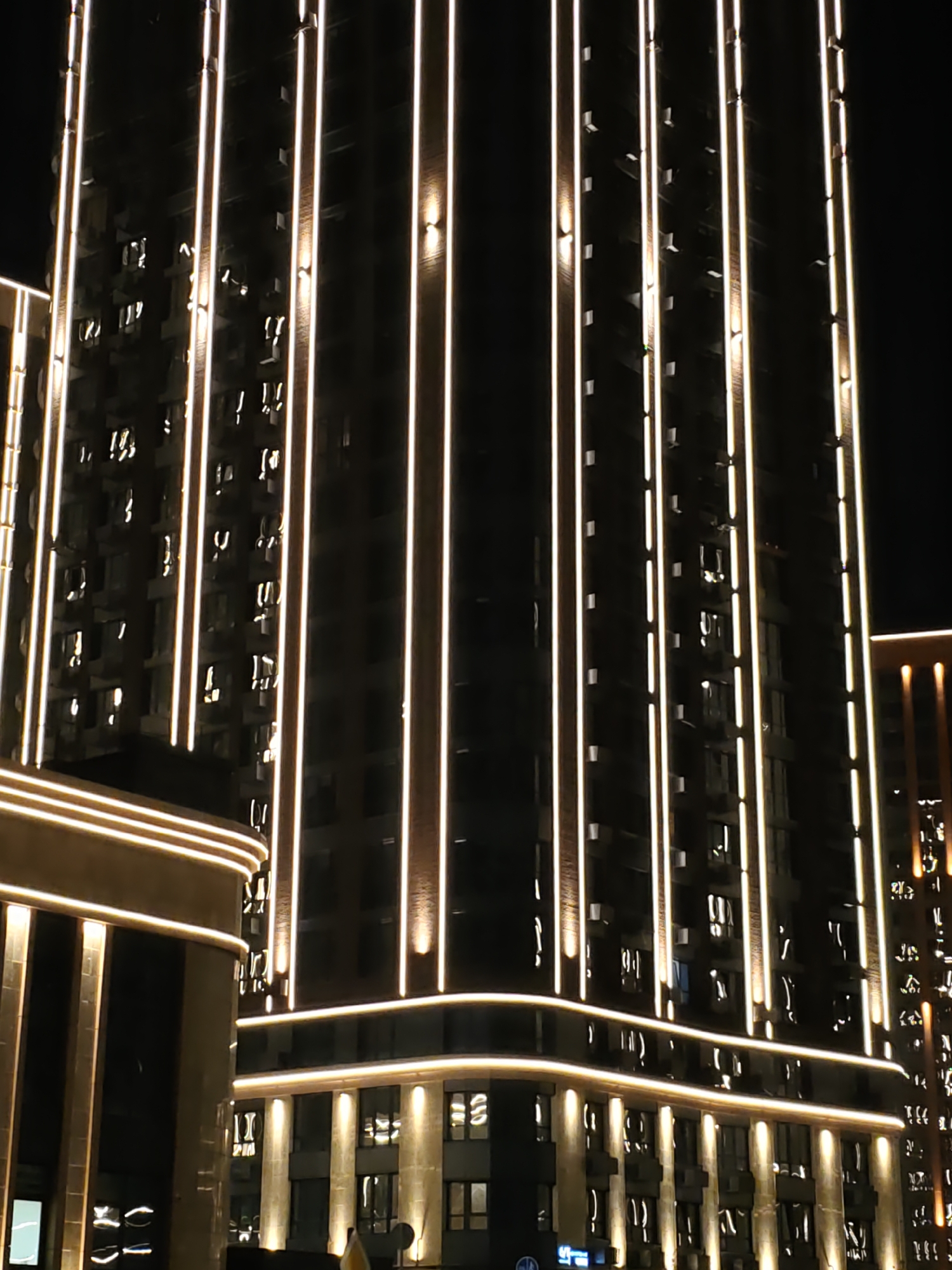 The image shows a modern high-rise building illuminated by vertical strips of warm white LED lights at night in Rostov-on-Don, Russia. The building's facade is dark, contrasting with the bright lights that highlight its architectural features. Windows are visible on the building, some with interior lights on, suggesting occupancy. In the foreground, a lower, curved building also features similar lighting. The overall scene depicts an urban nightscape with contemporary architecture.