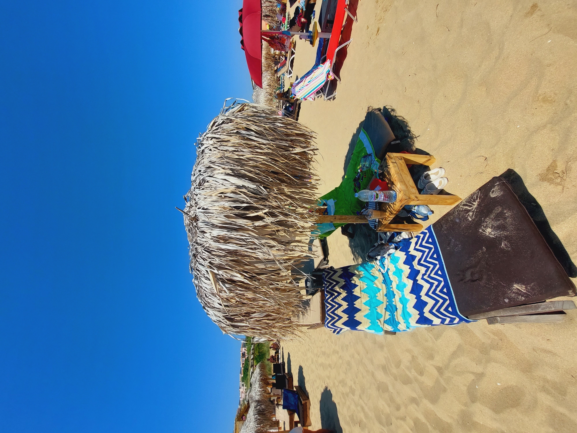 This is a photograph of a beach in Sozopol, Bulgaria, on a clear, sunny day. In the foreground, a wooden sun lounger is covered with a patterned towel featuring blue, white, and beige zigzags. To the right of the lounger, a small wooden table holds a bottle of water, a coffee cup, and a red cap. Two pairs of white sandals are placed underneath the table. A large thatched umbrella provides shade for the lounger and table. In the middle ground, more sun loungers and umbrellas are visible, some occupied by people. A red umbrella is prominent on the right side of the frame, with beach bags hanging from it. In the background, buildings are visible on a hillside. The sand is light brown and appears slightly damp in some areas. The sky is a clear, vibrant blue.