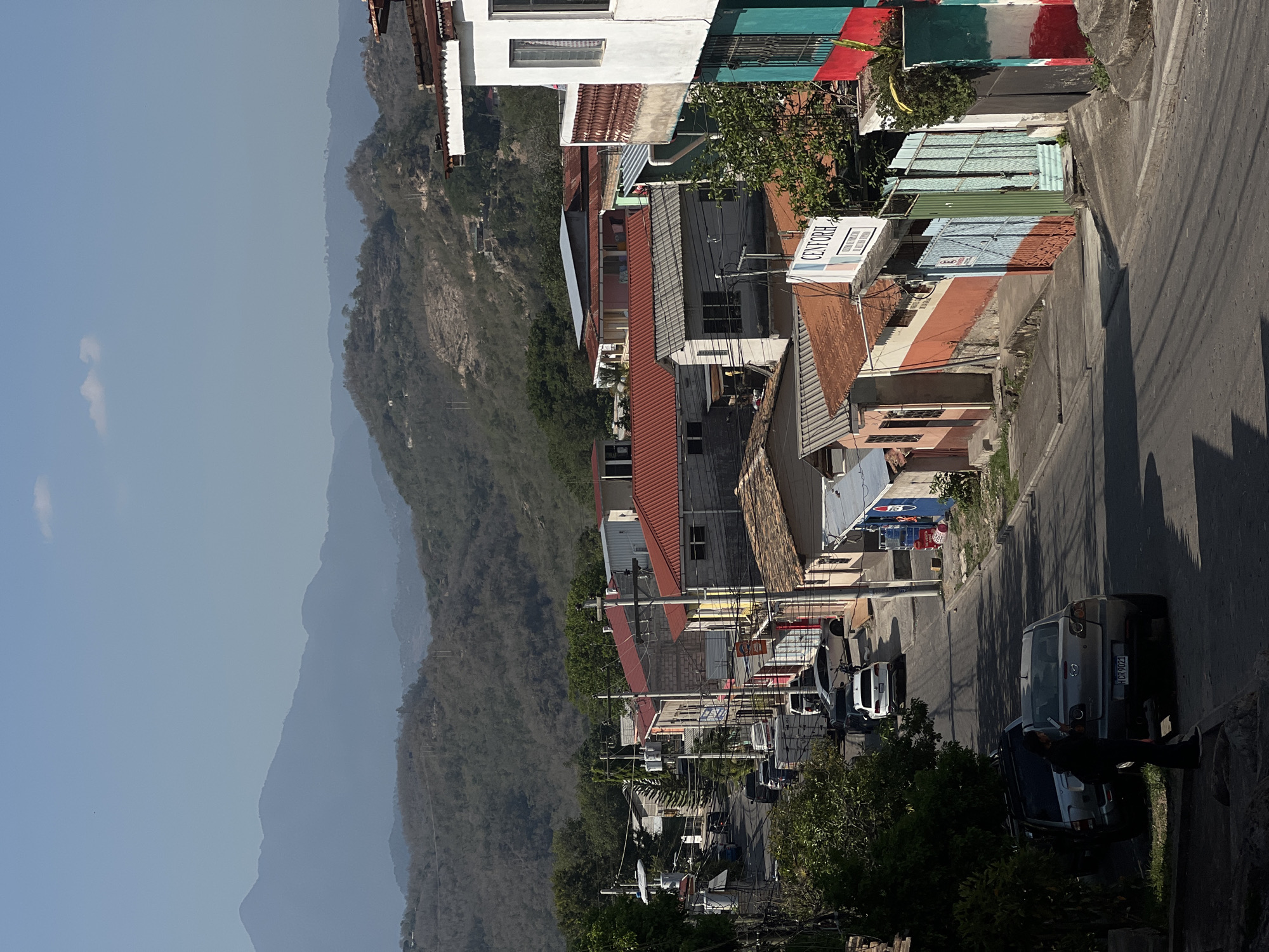 The image shows a street scene in Gracias, Honduras, with mountains visible in the background. A mix of residential and commercial buildings lines the street, some with colorful paint. Cars are parked along the street, and a person is standing next to one of the vehicles. Overhead power lines are strung between poles along the street. The mountains in the distance are covered in dense vegetation.