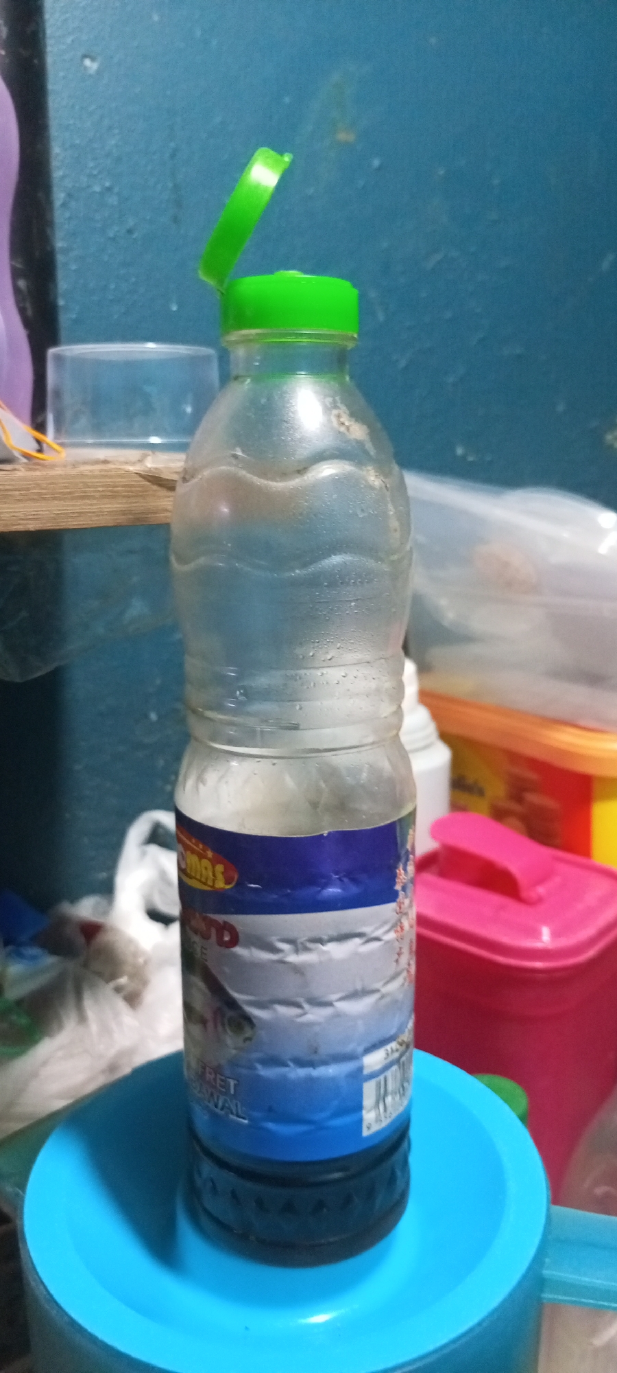 A clear plastic bottle with a green flip-top cap is positioned in the center of the frame. The bottle contains a clear liquid and has a label that features blue and white bands, with an image of a fish visible on the lower band. The bottle is placed on a bright blue, shallow circular container. In the background, a textured blue wall is visible, with various household items such as a pink plastic container with a lid, a yellow and orange container, and a white plastic bag with contents. A wooden shelf is partially visible on the left side of the image. The scene appears to be set indoors, possibly in a kitchen or storage area. There is no indication of the specific location in Kampung Telaga Air, Malaysia, beyond it being a domestic environment.