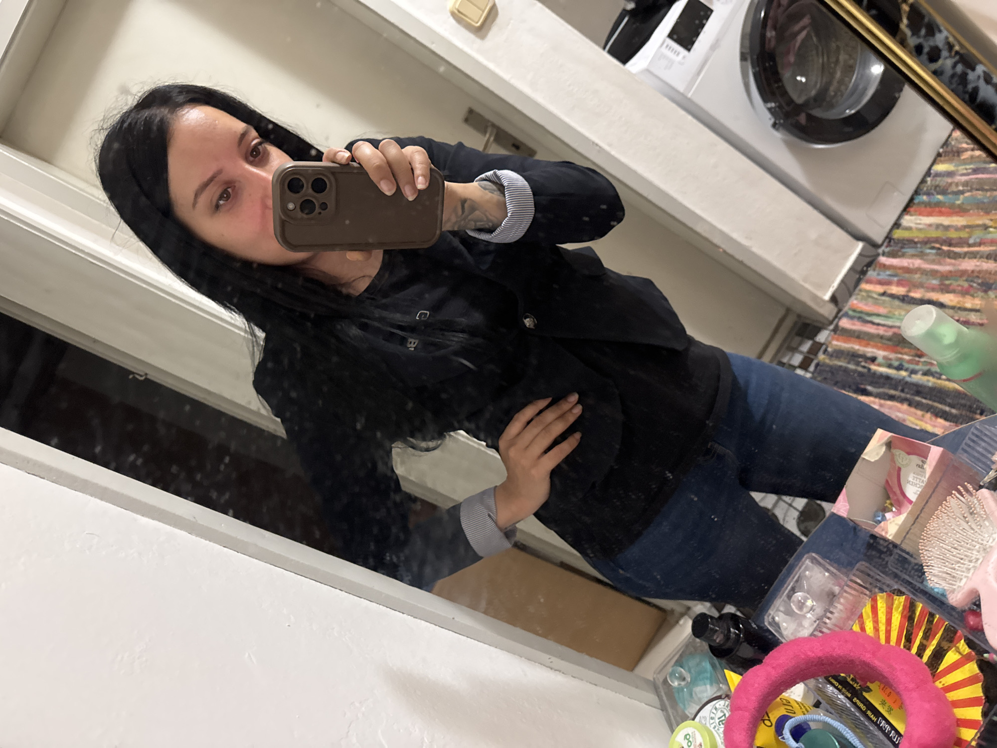 An individual, appearing female, with long dark hair, is depicted in a mirror selfie taken indoors in a room in the city of Unknown, Unknown. The subject wears a black long-sleeved top, a dark navy or black blazer with the cuffs folded to reveal blue and white stripes, and blue jeans. A tattoo is visible on the subject's left forearm. The right hand holds a brown-cased smartphone, positioned to capture the reflection, while the left hand rests on the subject's hip.
The background, reflected in a large, white-framed mirror, shows a domestic interior. A white doorway is visible behind the subject to the left. The floor is covered by a multi-colored striped rag rug. To the right, a white front-loading washing machine is present, alongside a light-colored wall-mounted fixture. In the foreground, on a surface below the subject's waist, various items are arranged, including a pink fuzzy headband, multiple clear plastic organizers containing small objects, a white hairbrush, a small black spray bottle, and a green spray bottle. A golden-framed object is partially visible on the far right of this surface. The lighting is consistent with indoor artificial illumination.