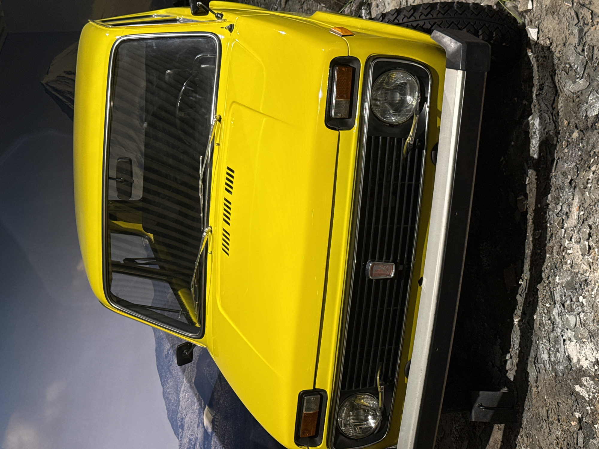 A close-up shot, taken from an elevated angle, reveals the front portion of a yellow Lada Niva, prominently displaying its grill, headlights, and bumper. The car is situated in what appears to be a museum setting, with a backdrop showing a mountain scene, while the ground around the car mimics a rough terrain, possibly to reflect the car's off-road capabilities. The image was taken at the Museum of Military Equipment UMMC, located in Verkhnyaya Pyshma, Russia.