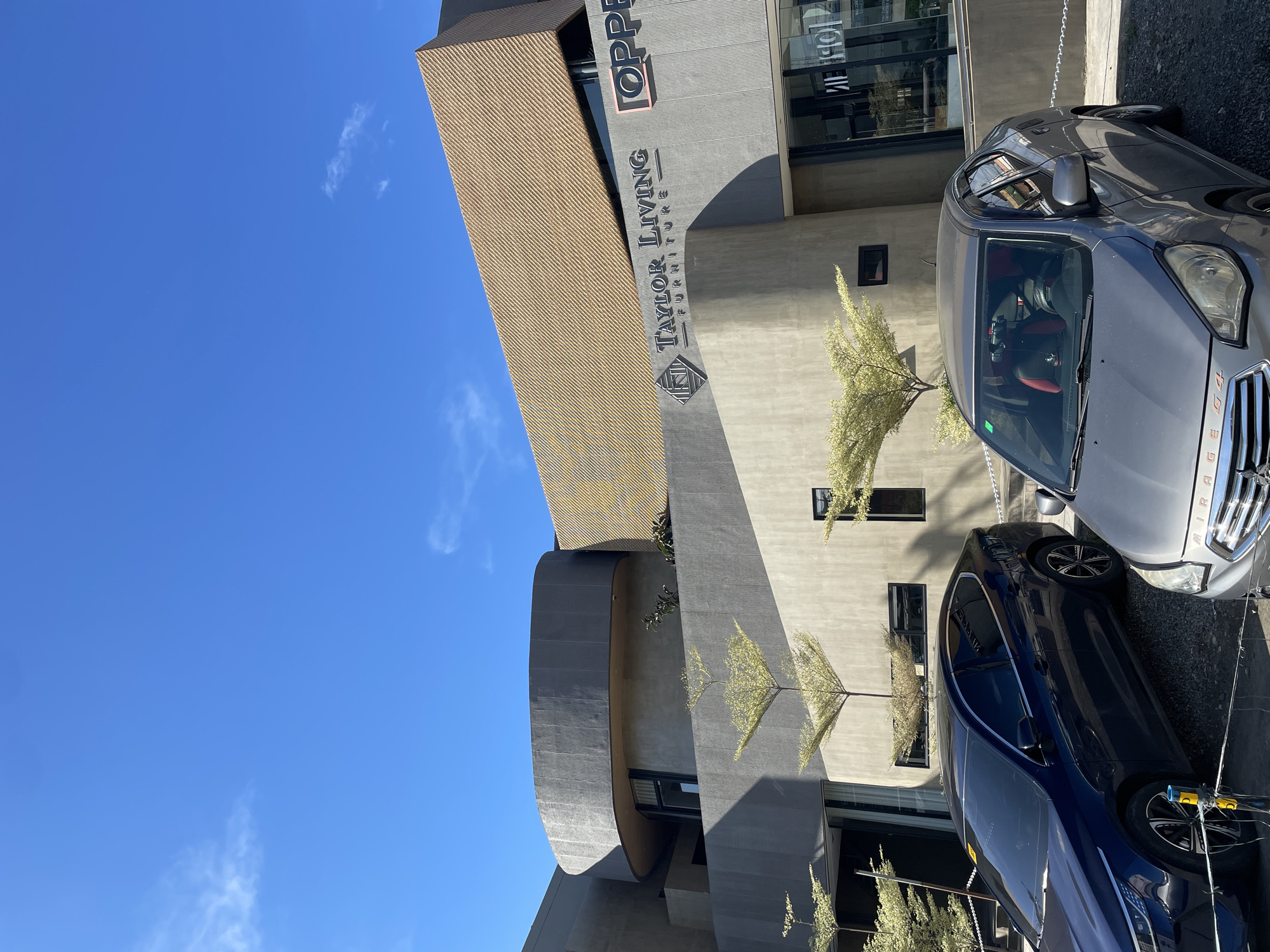 The image shows the exterior of a modern building with a distinctive architectural design, featuring curved elements and a textured golden facade. A sign on the building reads "TAYLOR LIVING FURNITURE". In the foreground, two cars are parked: a dark blue sedan on the left and a gray sedan on the right. The sky is clear and blue. The location appears to be a commercial area in Davao City, Philippines, specifically the building for Taylor Living Furniture, a known establishment in the city. The scene is well-lit by sunlight, suggesting it was taken during the day. There are no people visible in the image.