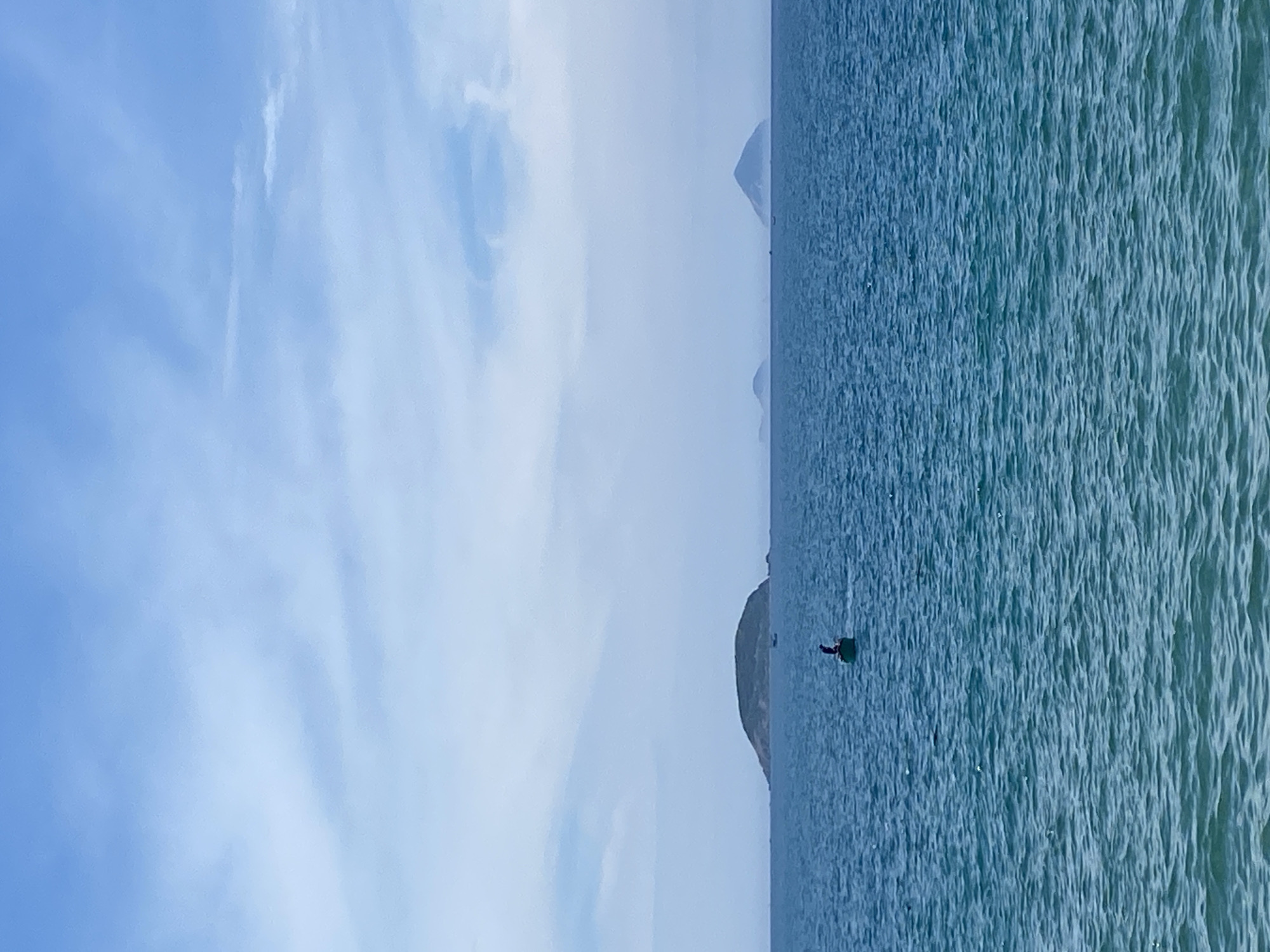 A wide shot captures the expanse of the ocean off the coast of Nha Trang, Vietnam, under a hazy sky. The water, a deep blue with a greenish hue, is rippled by a gentle breeze. In the foreground, the ocean dominates the frame, with subtle variations in color indicating depth and movement. In the mid-ground, a solitary figure is seated in a small, dark-hulled boat, positioned slightly off-center to the left. The boat appears to be a traditional fishing vessel. Further in the distance, a chain of islands stretches across the horizon, their outlines softened by atmospheric haze. The largest island, a lush green mound, is the most prominent, with a few smaller islands dotting the seascape behind it. The sky above is a pale blue, streaked with wispy white clouds, suggesting a slightly overcast but calm day. The overall impression is one of tranquility and a vast, open seascape.