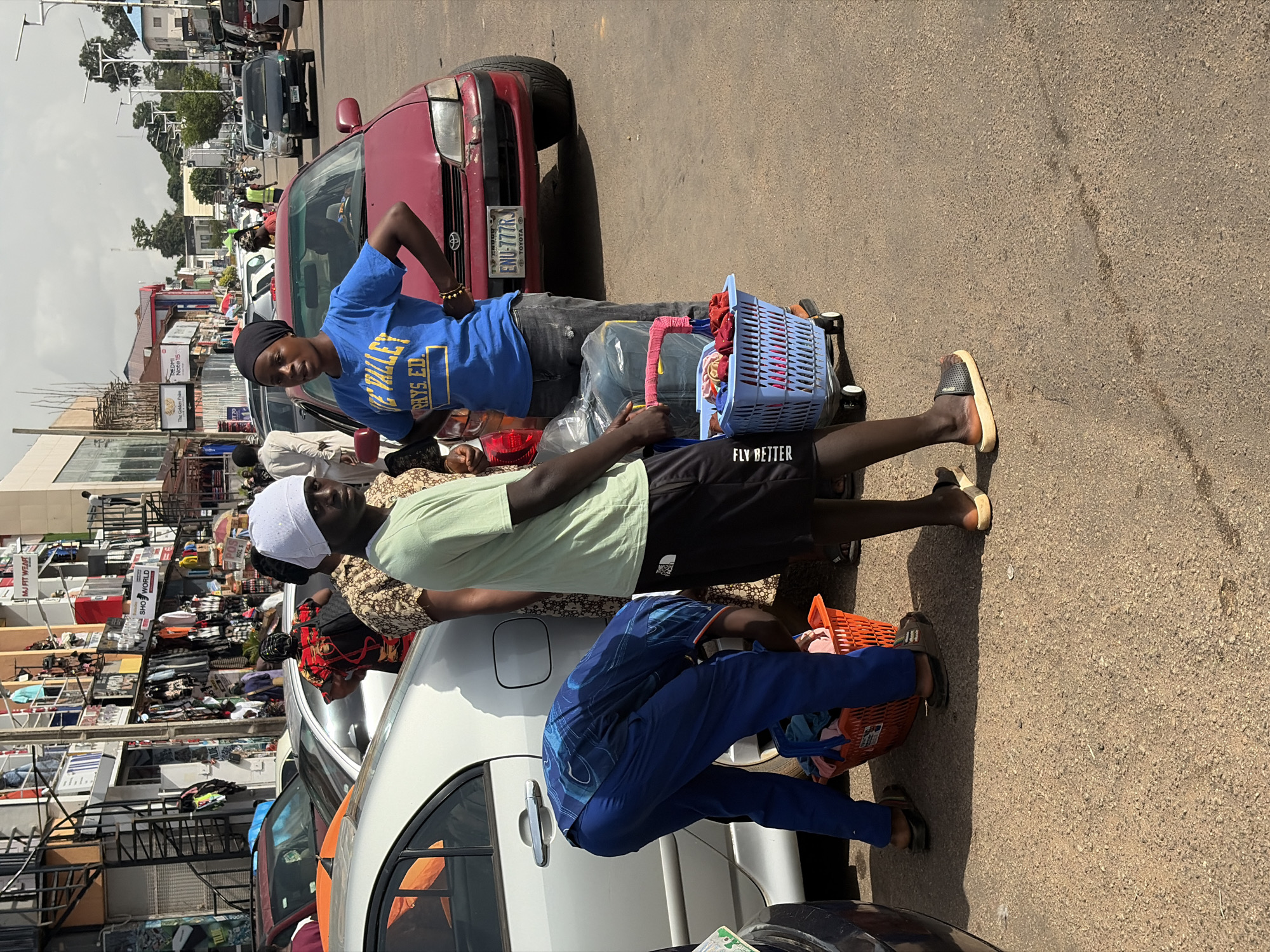 The image depicts a street scene in Jos, Nigeria. In the foreground, two individuals are standing on a paved road. One is a young man wearing a light green t-shirt and black shorts, facing partially away from the camera. He appears to be holding a blue plastic basket with wheels, filled with various items. Next to him, a young woman is wearing a blue t-shirt with yellow lettering and dark jeans. She is looking towards the camera, with her hands on her hips. Partially visible behind them is a red car. To the left, a person in blue pants and a blue patterned shirt is bent over, placing items into an orange plastic basket. Further back, several cars and motorcycles are visible, as well as buildings lining the street with shops displaying goods. The overall environment suggests a busy commercial area in an urban setting.