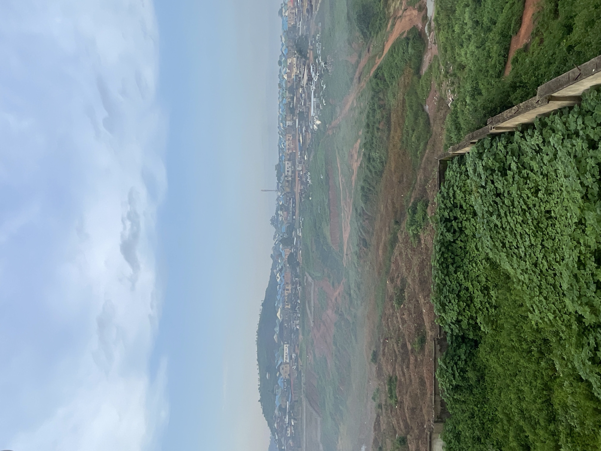 The image depicts a hazy, elevated view of a residential area on the outskirts of Abuja, Nigeria. In the foreground, a dense expanse of vibrant green foliage, possibly a cultivated area or overgrowth, dominates the lower portion of the frame. A low concrete wall, weathered and showing signs of age, runs horizontally across the middle ground, separating the dense greenery from the terrain beyond.
Beyond the wall, the landscape transitions to a more rugged and uneven terrain, characterized by exposed reddish-brown soil and patches of sparse vegetation. This area appears to be undergoing some form of development or landscaping, with visible earthworks and tiered slopes. A dirt road winds its way through this section.
Further in the distance, a sprawling settlement is visible, characterized by numerous buildings with colorful roofs, predominantly blue and yellow. The houses are densely packed, indicating a medium to high-density residential area. A prominent hill rises in the background, also dotted with structures, suggesting the settlement extends across varied topography. The overall atmosphere is subdued due to a light haze or mist, which softens the details of the distant cityscape and creates a sense of depth. Several vehicles are scattered throughout the settlement and on the roads, suggesting activity within the community. The sky above is overcast with a pale blue and grey hue, consistent with early morning or a cloudy day.