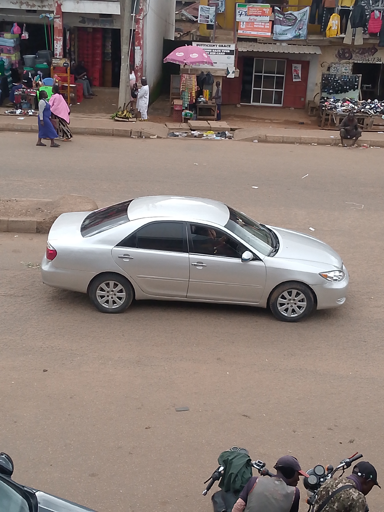 In Bukuru, Nigeria, a silver Toyota Camry sedan is parked on a paved road. The car is silver with tinted windows and appears to be a 2007-2011 model. The left rear tire is flat. In the background, a street market is visible, with stalls displaying various goods, including fabrics, shoes, and food items. Several people are present, some browsing or working at the stalls, while others are walking along the sidewalk. A group of individuals are gathered near a motorcycle in the foreground, with one person appearing to adjust a green bag on the back of the motorcycle. The environment is characteristic of a bustling urban market scene in Nigeria.