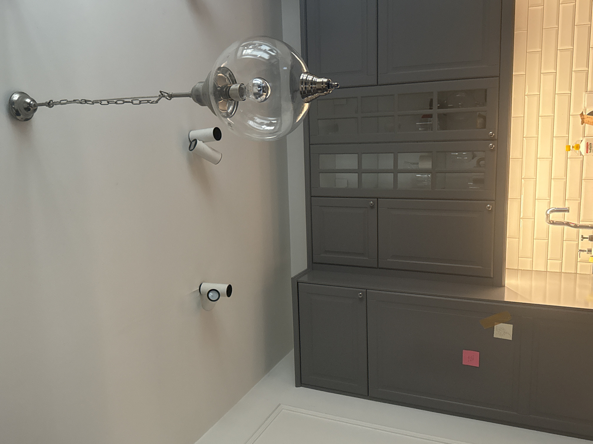 The image displays a modern kitchen interior, oriented vertically. In the foreground, a sleek pendant light with a glass globe and a silver chain hangs from the ceiling. Adjacent to the light, two small white security cameras are mounted on the wall. The kitchen features dark gray cabinets, some with glass fronts revealing contents, and a smooth countertop. A tiled backsplash in a light color runs along the wall behind the countertop, illuminated by warm lighting. Two colorful sticky notes, one pink and one yellow, are attached to the cabinet doors in the lower right section of the image. The scene is indoors, and the lighting suggests it could be daytime, possibly artificial light. No people are visible, and the overall atmosphere is clean and organized.