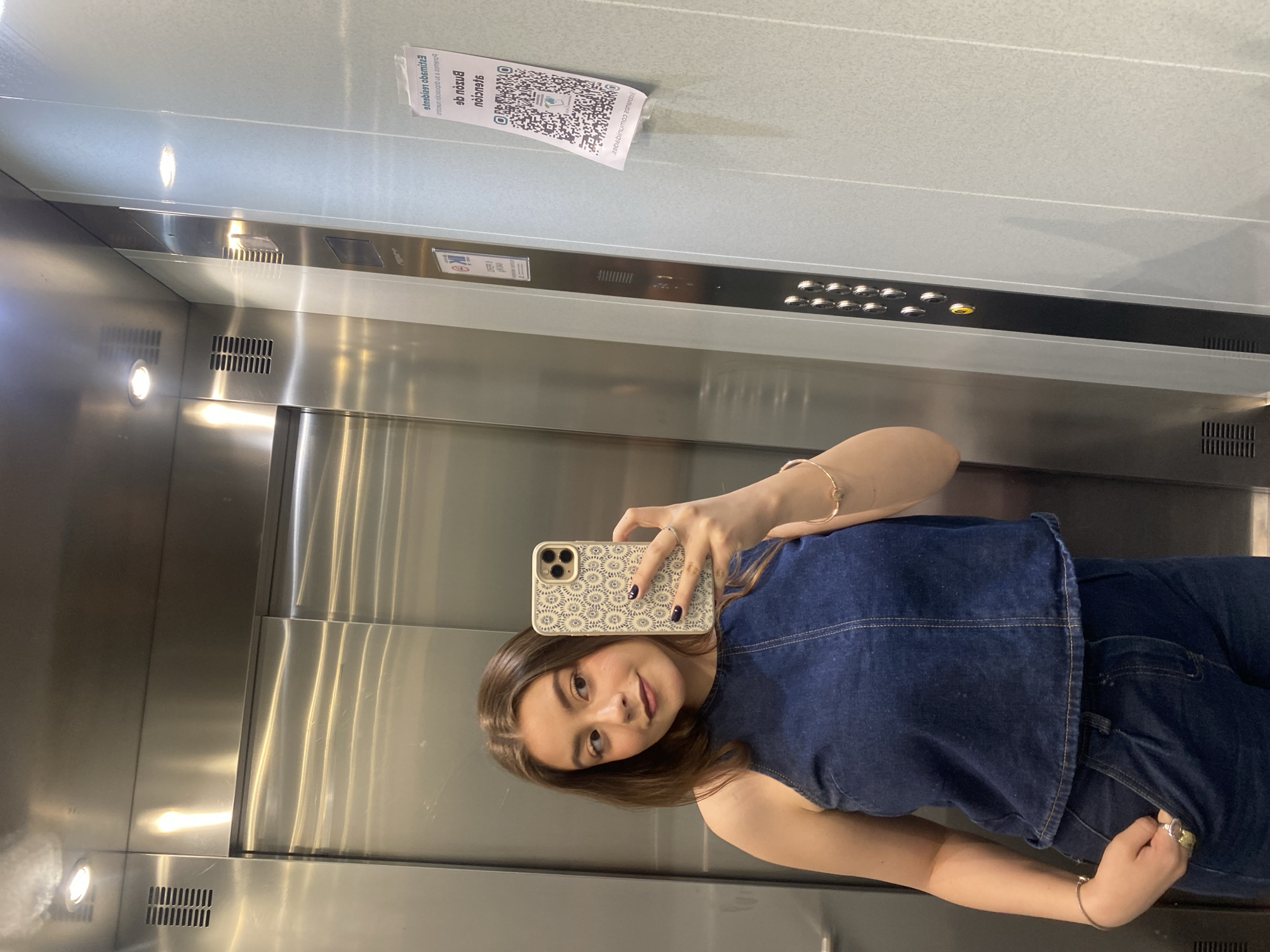 A young woman is taking a selfie in an elevator. She is wearing a dark blue denim top and jeans. She has brown hair and is holding her phone up to take the picture. The elevator is made of stainless steel. There is a QR code on the wall of the elevator. The location is Unknown, Unknown.