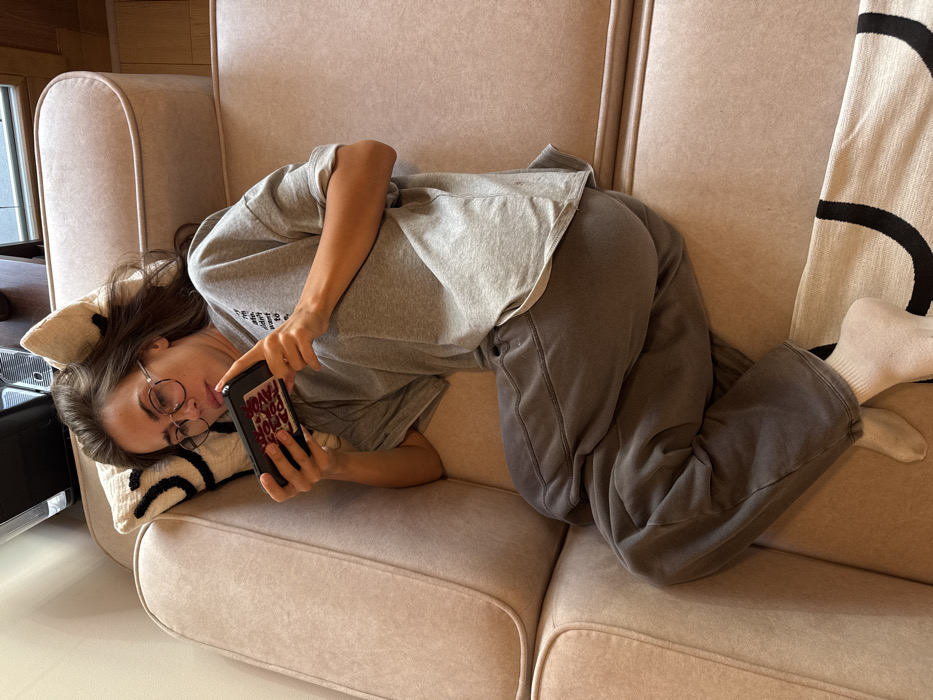 A young adult female is observed in a reclined position on a light beige upholstered sofa, situated indoors in Warsaw, Poland. She wears a grey oversized t-shirt, dark grey sweatpants, and white socks. Her head is supported by an off-white decorative pillow featuring black abstract designs, and she is wearing eyeglasses with dark frames. Both hands hold a black smartphone horizontally, the rear of which displays a white rectangular patch with partially visible red text reading "AVO" and "TOON". Her gaze is directed intently at the phone screen. A light-colored rug with black curved patterns is present on the floor beneath her feet. The background reveals a segment of a light-colored wall and the top portion of a dark, unspecified object resting on a surface.