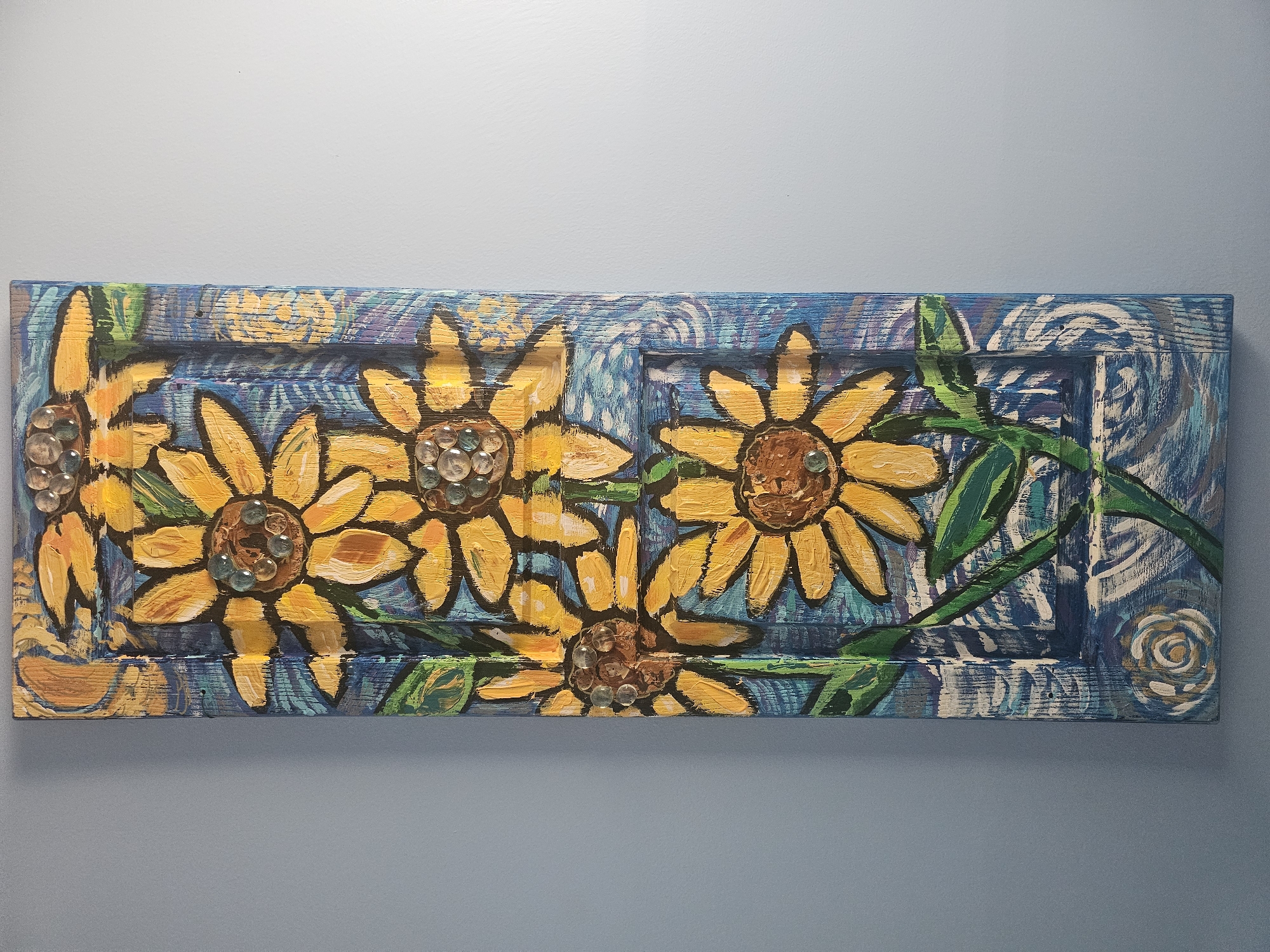 A vertical, rectangular painting featuring three sunflowers is mounted on a light blue wall. The painting is done on a textured surface, possibly wood, with visible grain and imperfections. The sunflowers are rendered in vibrant yellow and orange, with brown centers that are decorated with small, clear, round embellishments. Green leaves and stems are depicted in a somewhat abstract style. The background of the painting is a deep blue with swirling patterns in lighter blues, white, and hints of purple, giving a sense of movement. The overall style is folk art. There are no people present in the image. The painting is hanging in a room with neutral lighting. This scene was captured in Marietta, United States.