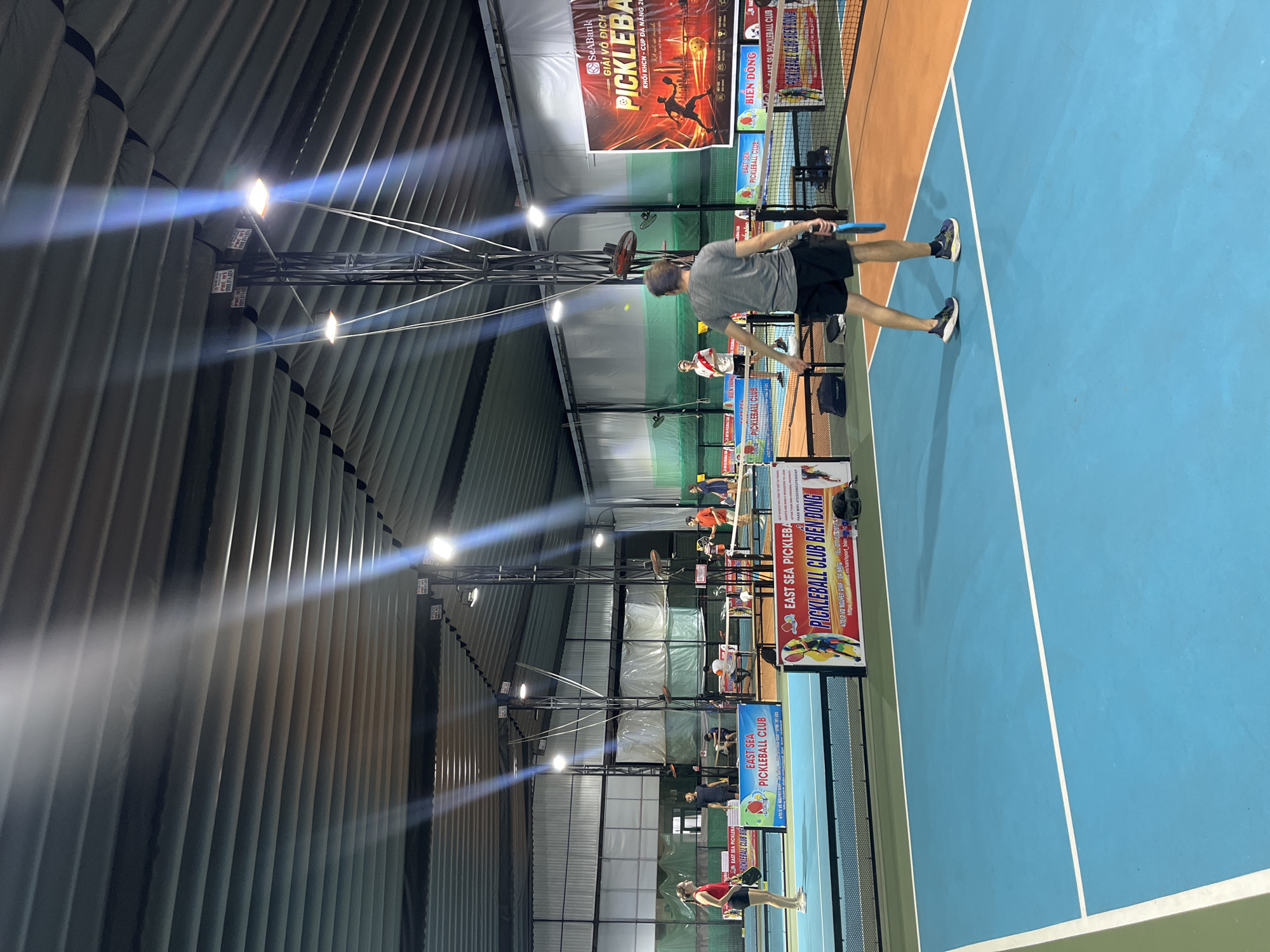 An indoor pickleball court in Da Nang, Vietnam, is captured from a low angle, showing players engaged in a game. The court features a blue playing surface with white lines and an orange outer boundary. A net divides the court. Several players are visible, including a woman in a red top and black shorts on the left, a man in a grey t-shirt and black shorts in the foreground holding a blue paddle, and other players on the far side of the court. The environment is well-lit by overhead fluorescent lights attached to metal trusses that run along the ribbed ceiling. Banners advertising "East Sea Pickleball Club Bien Dong" and a "SeaBank Pickleball Cup" are displayed around the court. The overall scene depicts an active sports facility.
