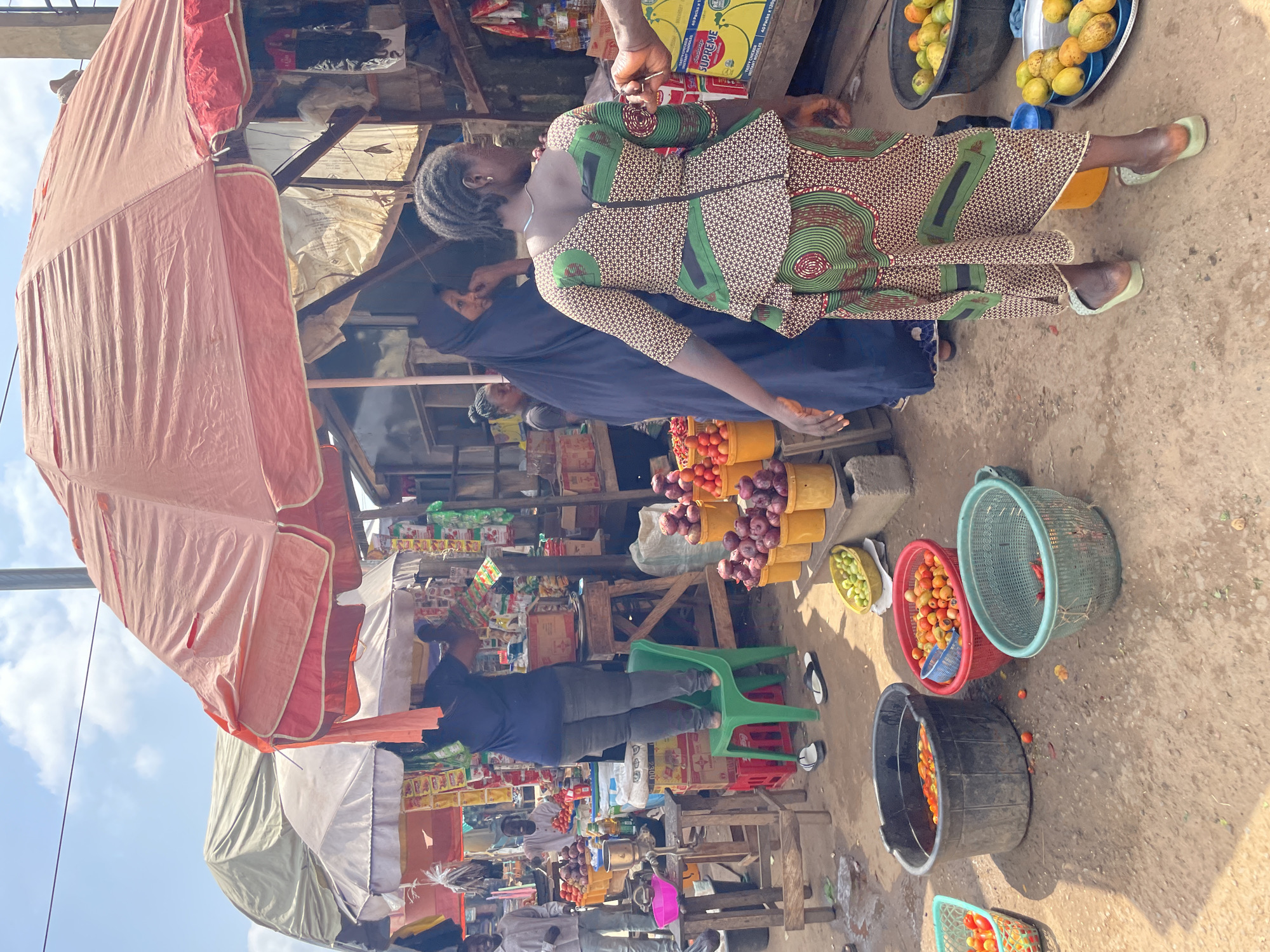 This image captures a vibrant market scene in Akwanga, Nigeria. The foreground is dominated by a variety of fruits and vegetables displayed in baskets and buckets. Several individuals are engaged in transactions or attending to their stalls.
In the center, a woman wearing a dark blue t-shirt and grey trousers stands on a green plastic chair, reaching towards goods suspended from an overhead canopy. To her right, another woman in a colorful patterned dress is turned away from the camera, facing a stall with an array of products. Behind them, a woman in a dark hijab is visible.
To the left, two men stand near stalls laden with produce. One man in a light-colored shirt smiles at the camera while another, partially obscured, appears to be working. Various stalls are set up under large, colorful umbrellas, offering a diverse selection of goods, including packaged items and fresh produce.
The ground is a dusty, uneven surface, typical of an outdoor market. The background reveals more stalls and structures, suggesting a densely packed commercial area. The sky above is partly cloudy, indicating a bright day. The overall impression is one of lively commerce and daily life in Akwanga.
