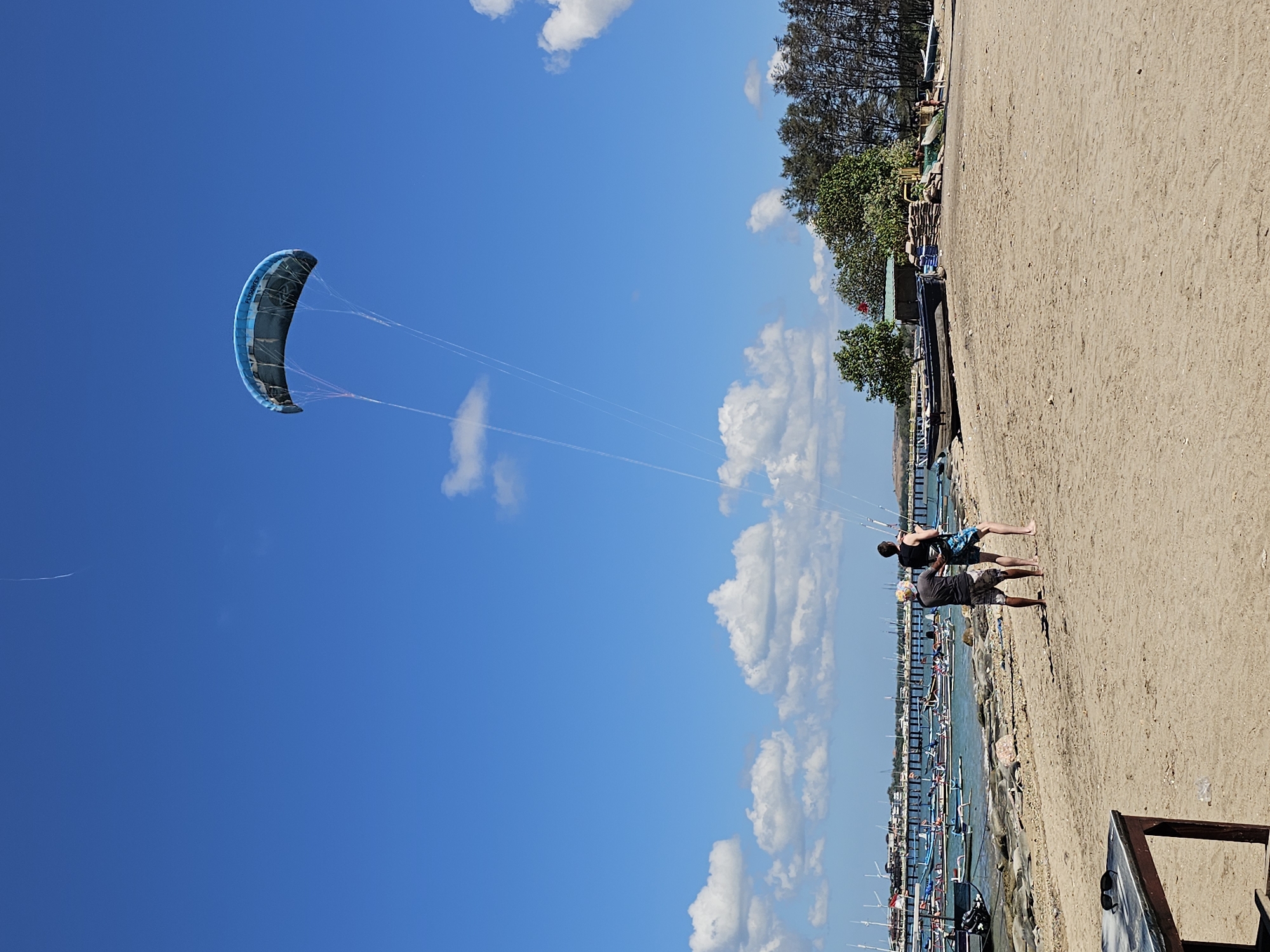 The image depicts a beach scene in Denpasar, Indonesia, with two individuals preparing to launch or control a large, blue kite. The kite is airborne, its strings taut and extending downwards towards the people on the sandy shore. The water is visible behind them, dotted with numerous traditional Indonesian fishing boats, some moored and others in the water. A long pier or jetty stretches out into the sea in the background, with buildings and trees lining the coastline. The sky is a vibrant blue, with scattered white clouds. The foreground features a sandy beach with some scattered debris and a low table with sunglasses on it. The overall atmosphere is one of outdoor recreation and coastal activity.