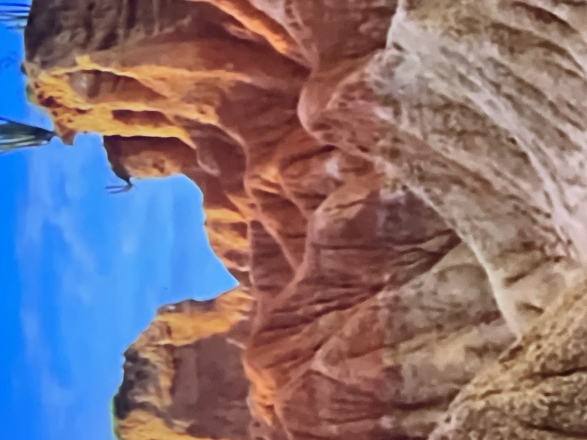 The image is a landscape of arid, eroded rock formations under a bright blue sky with scattered clouds. The formations are predominantly orange and brown hues, with some lighter, sandy colored areas. Jagged peaks and valleys create a rugged terrain. In the upper right portion of the image, a thin, green cactus-like plant is visible growing from the top of one of the formations. The scene is consistent with a desert or badlands environment. There is no indication of the city of Casablanca, Morocco in this image.