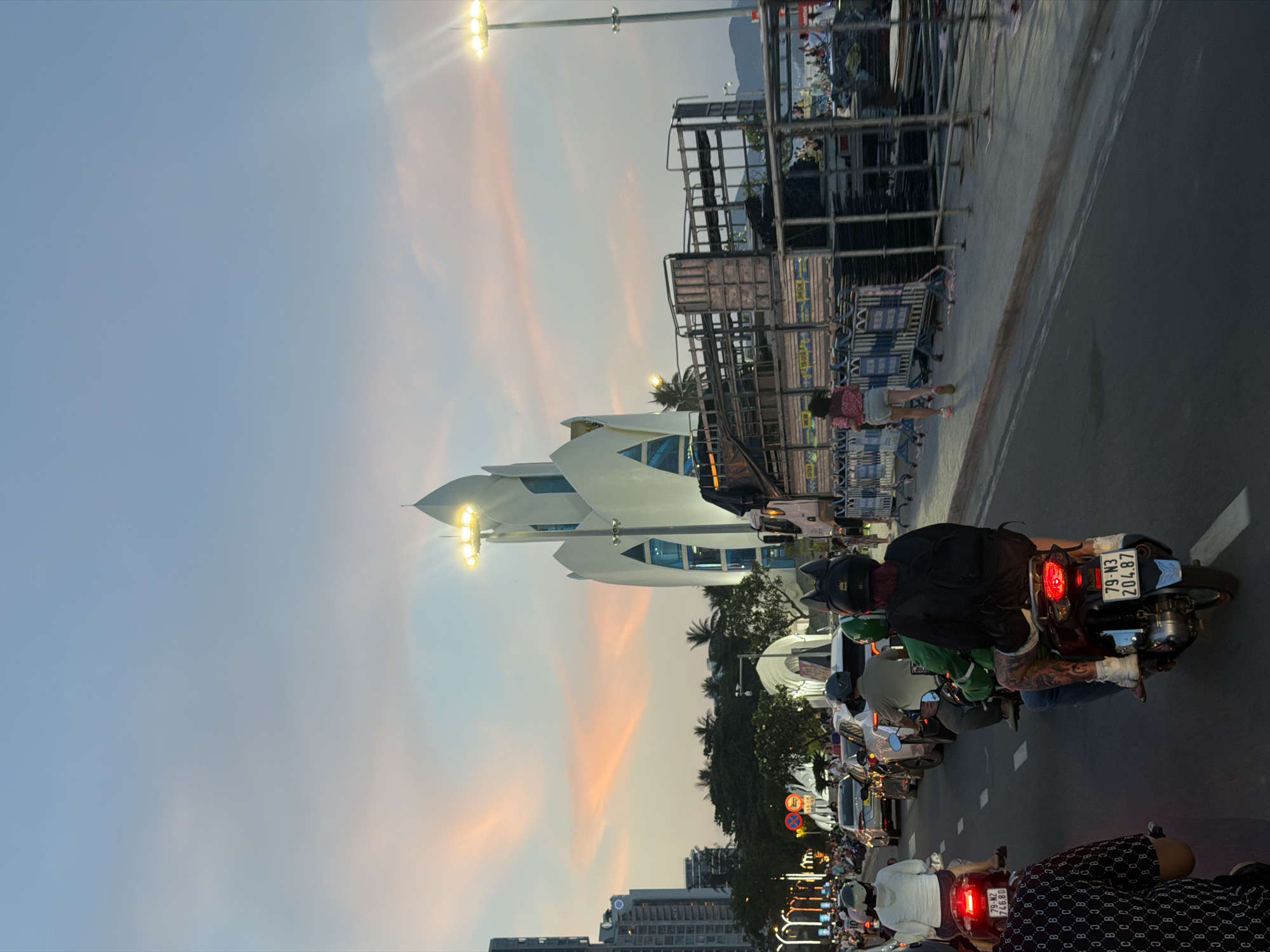 The image depicts a street scene in Nha Trang Ward, Vietnam, during dusk. A white, lotus-shaped building with illuminated windows is prominently featured on the right side of the frame. In front of the building, scaffolding and construction materials are visible. A street lamp with multiple bulbs illuminates the area. The sky is a gradient of blue and pink, indicating sunset.
The street is filled with traffic, primarily motorcycles. Several individuals are riding motorcycles, some with passengers. One motorcycle in the foreground has a license plate reading "79-N3 204.87." People are dressed in casual clothing. Buildings and palm trees line the street, suggesting an urban coastal environment. The overall atmosphere is one of a bustling city at the end of the day.