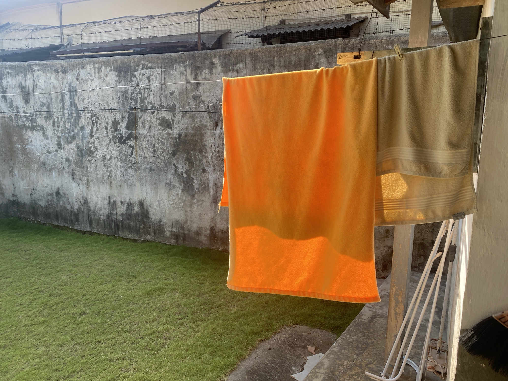 In Kotakulon, Indonesia, two towels are hanging on a clothesline in a backyard. One towel is bright orange and the other is beige. A weathered concrete wall with a barbed wire fence along the top is behind the clothesline. In the foreground, a patch of green grass is visible. The scene is illuminated by the warm light of the sun.