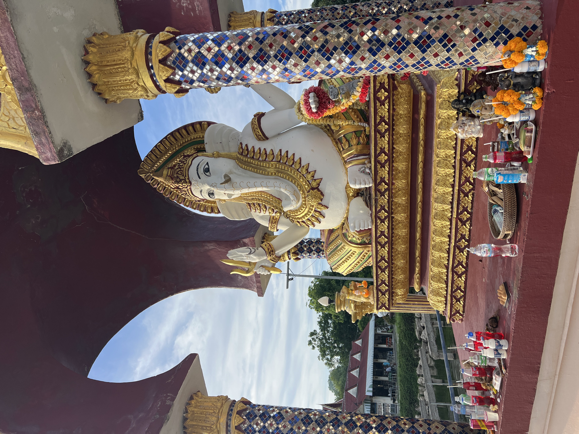 The image shows a large statue of Ganesha, the Hindu deity, seated on a golden platform. The statue is white with ornate gold embellishments. It is situated in a shrine or temple-like structure with red arched coverings and decorative pillars. The pillars are covered in colorful mosaic tiles. In front of the shrine, on a red ledge, are offerings including bottles of drinks, food items, and smaller statues, some adorned with marigold garlands. In the background, there is a body of water, trees, and a building with a red roof and awnings, suggesting a tropical or subtropical environment. The sky above is blue with scattered clouds. The location is identified as Baan Plai Laem, Thailand.