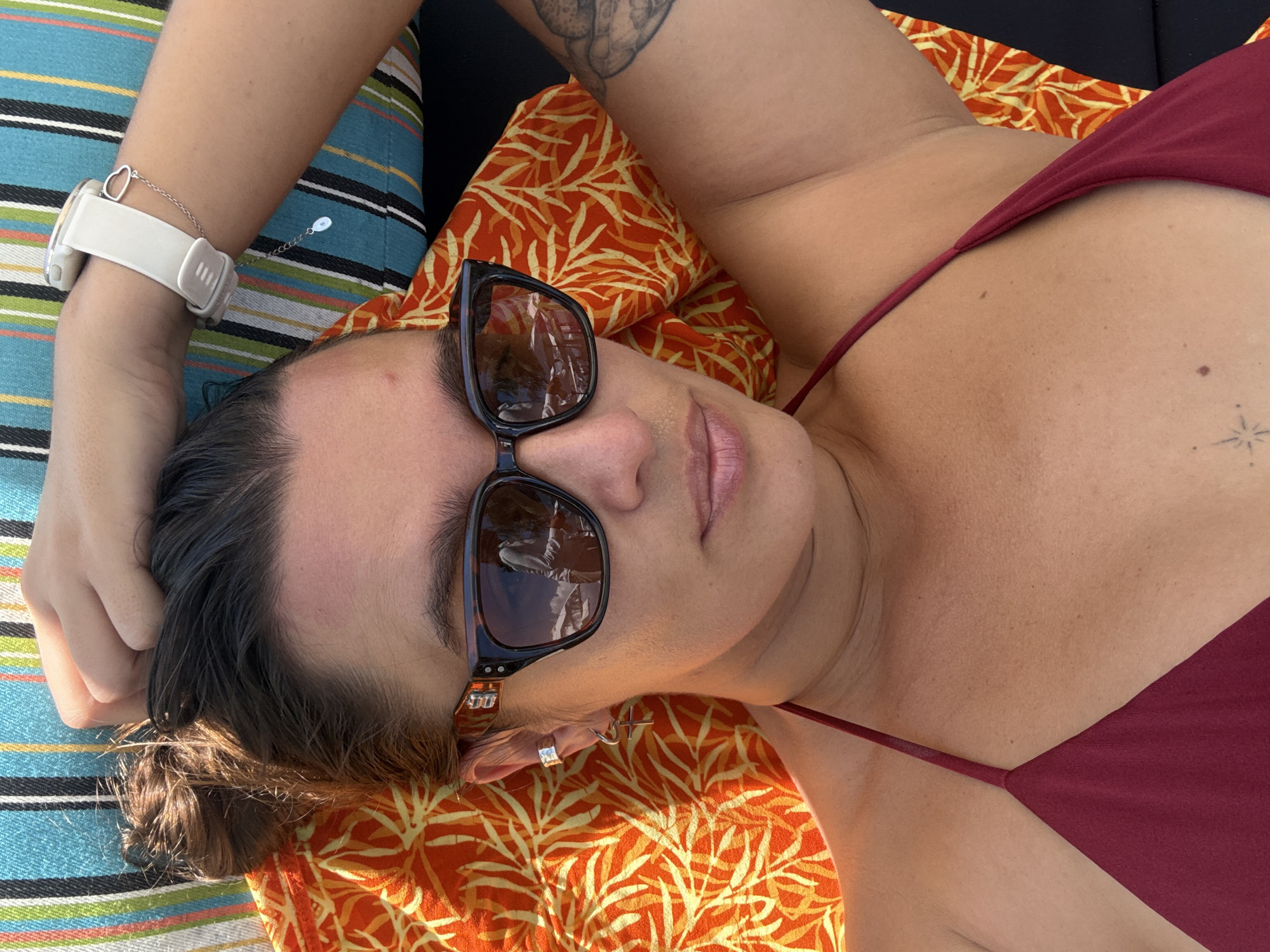 A woman is reclining, likely outdoors, wearing sunglasses and a maroon-colored top. She has fair skin, dark hair pulled back, and wears a white smartwatch on her left wrist. Her right arm is raised with a tattoo visible on her inner arm. She is lying on a striped cushion with an orange and yellow patterned pillow underneath. The location cannot be determined from the image.