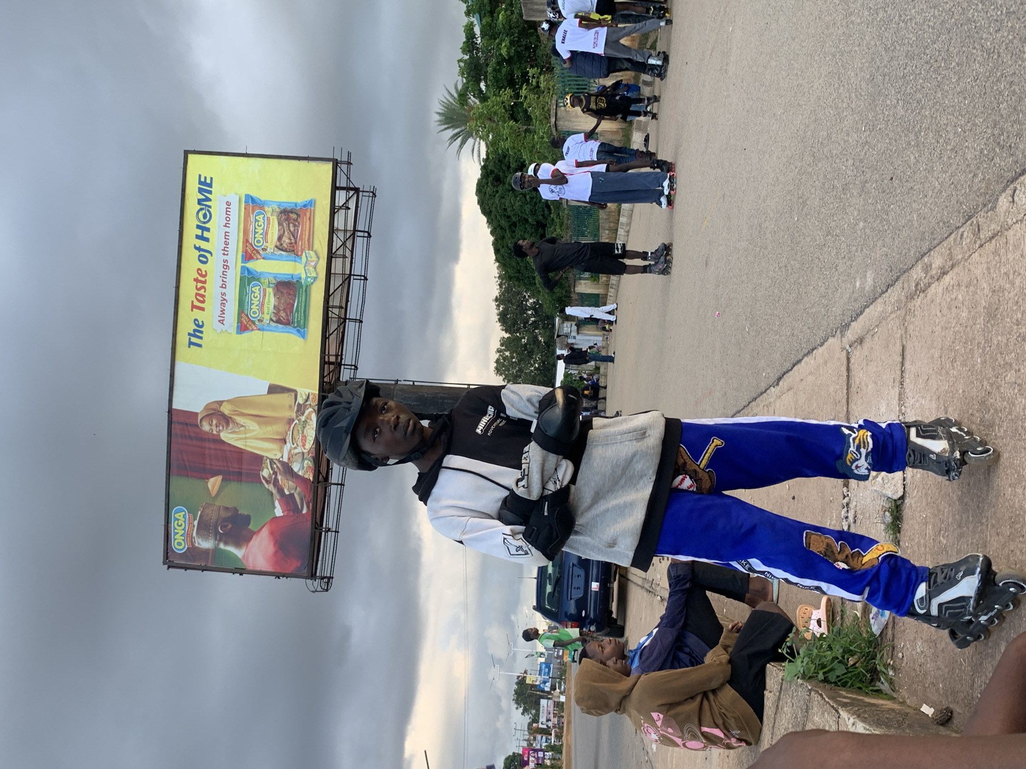 The image captures a street scene in Jos, Nigeria, under an overcast sky. In the foreground, a young person wearing a helmet, black and white hoodie, and blue pants with cartoon patches stands on rollerblades, facing the camera with arms crossed. To the left, two individuals sit on a concrete ledge, one in a brown hooded sweatshirt and the other in a blue jacket. In the background, several other people on rollerblades are visible on the road. A blue SUV is parked on the side of the road. Dominating the upper portion of the image is a large billboard advertising "ONGA" food seasoning. The billboard features images of a man and a woman sharing a meal, and packets of "ONGA" seasoning, with the tagline "The Taste of HOME" and "Always brings them home." Trees and other buildings are visible further in the background. The overall impression is that of a casual gathering of people engaged in recreational activity on a public street.