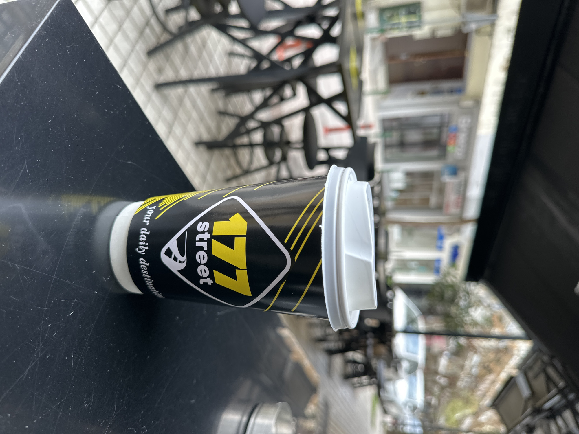 A coffee cup from "177 street," a business located in Serres, Greece, sits on a dark-colored table outdoors. The cup is primarily black with a yellow logo and a white lid. The logo displays "177 street" in yellow text inside a white and black geometric shape. Text on the cup reads "Your daily destination." The table surface is glossy and shows some scratches. The background features an outdoor urban setting, including other tables and chairs, and a view of buildings across the street. The chairs have a black frame with a complex geometric pattern. The buildings show multiple levels, windows, and signs, indicating an active commercial area.