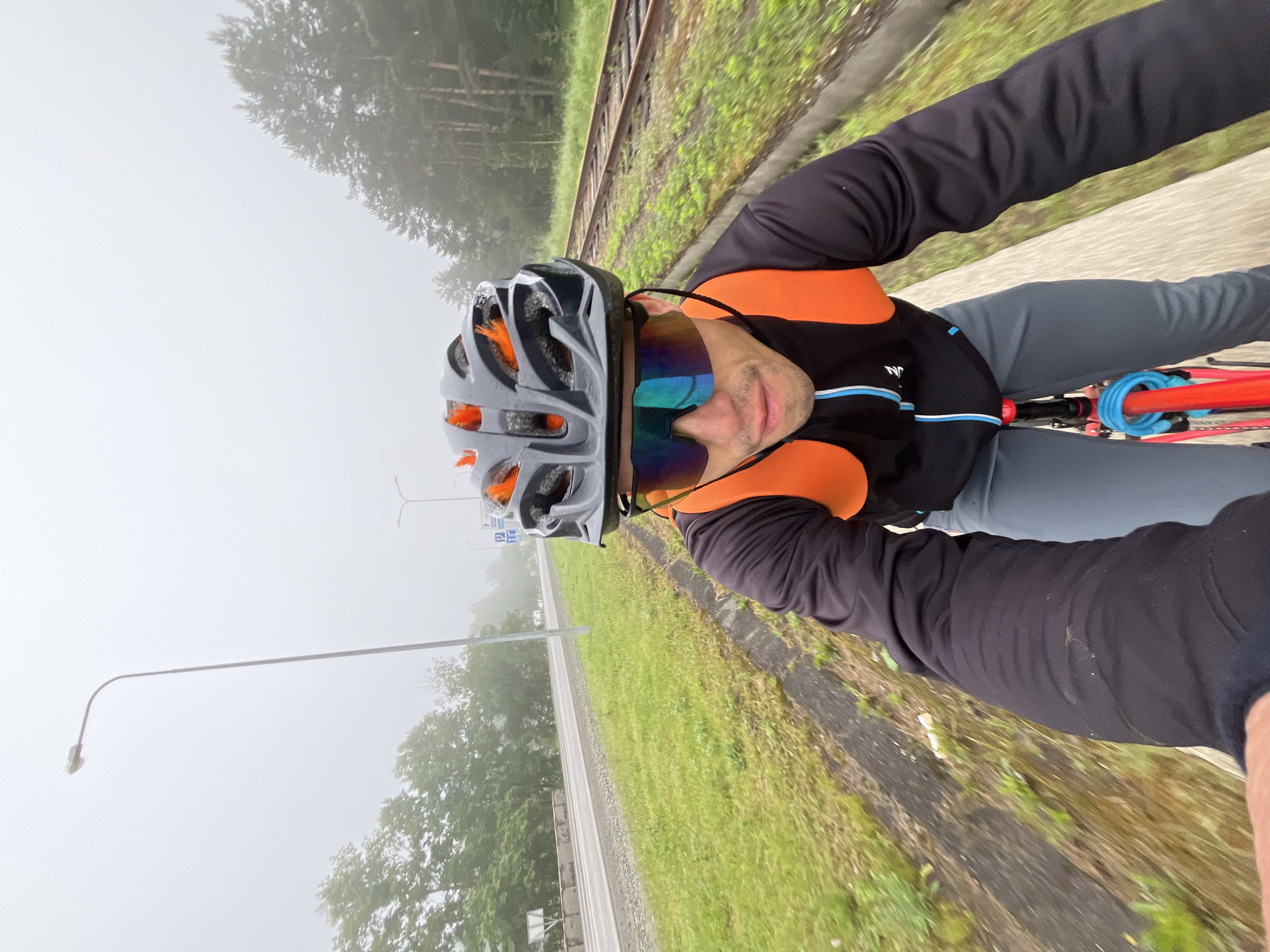 This image captures a first-person perspective selfie of a cyclist riding in Chaikovsky, Russia, on a heavily misty or foggy day. The composition is rotated, creating a diagonal horizon.
The primary subject is an individual, likely male, positioned from the chest upwards and facing the camera. He is equipped with cycling gear, including a grey and black helmet with distinct orange accents, and mirrored sunglasses that display a vibrant, multi-colored reflection. His attire consists of a black, long-sleeved cycling jersey featuring prominent orange sections on the shoulders and sides, and horizontal blue and white stripes across the chest. The letters "NYN" are discernible on the left side of his jersey. He wears grey cycling pants, and a segment of a red bicycle frame is visible, adorned with a bright blue coiled bike lock.
The outdoor scene is enveloped in thick mist or fog, resulting in diffused lighting and significantly reduced visibility, particularly in the background. This atmospheric condition lends a soft, muted quality to the landscape. To the right of the cyclist, a set of railway tracks runs parallel to the direction of travel, flanked by green grass. Beyond the tracks, trees are partially obscured by the haze. On the left side, a paved road or path is visible, bordered by more greenery, and a tall lamppost extends into the misty distance. The overall impression is one of a serene and somewhat secluded journey through an ethereal, fog-laden environment.