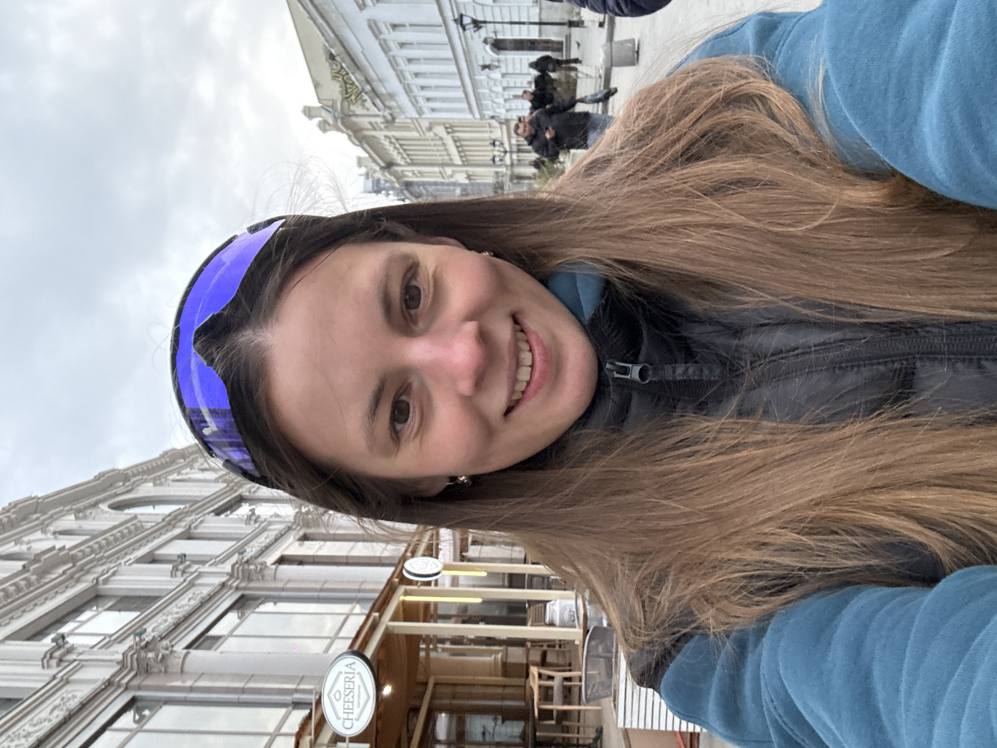 The image is a selfie, rotated 90 degrees clockwise, featuring a young woman smiling broadly at the camera. She has long, light brown hair and wears a blue long-sleeved top under a dark vest. Purple/blue mirrored sunglasses are pushed up onto her forehead. Her expression is cheerful.
The background shows a bustling city street in Kazan, Russia, lined with ornate, light-colored historical buildings. These multi-story structures feature numerous windows and decorative architectural details, such as columns and cornices. Several blurred figures of people are visible in the distance along the street, indicating pedestrian activity. The sky above is overcast and bright, suggesting a cloudy daytime setting.
A notable detail in the background is a circular sign affixed to one of the buildings, clearly displaying the word "CHEESERIA" in white text on a dark background, accompanied by a small graphic. The overall scene depicts an urban environment with classic European-style architecture, captured with a personal, direct gaze from the subject.