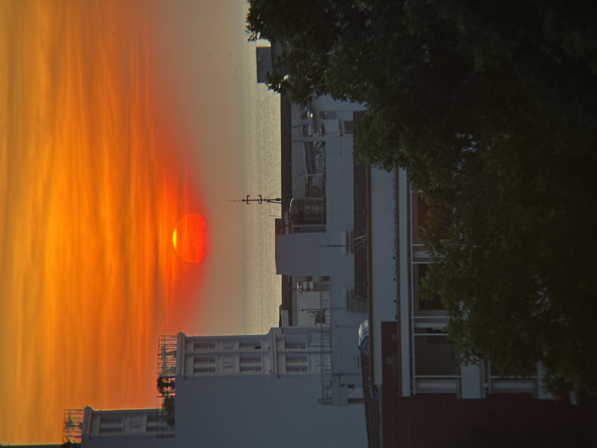 The image captures a vibrant sunset over the city of Nha Trang, Vietnam. The sky is a dramatic display of orange and red hues, with the sun a bright red orb peeking just above the horizon. In the foreground, several buildings are visible, their architecture suggesting a coastal urban setting. The tallest building on the left is white with multiple windows and decorative railings on balconies. Further back, other buildings are partially obscured by the intense light of the sunset and trees. The sea is visible in the middle ground, a muted gray expanse reflecting the colors of the sky. The overall scene is one of natural beauty meeting urban development during the twilight hours.