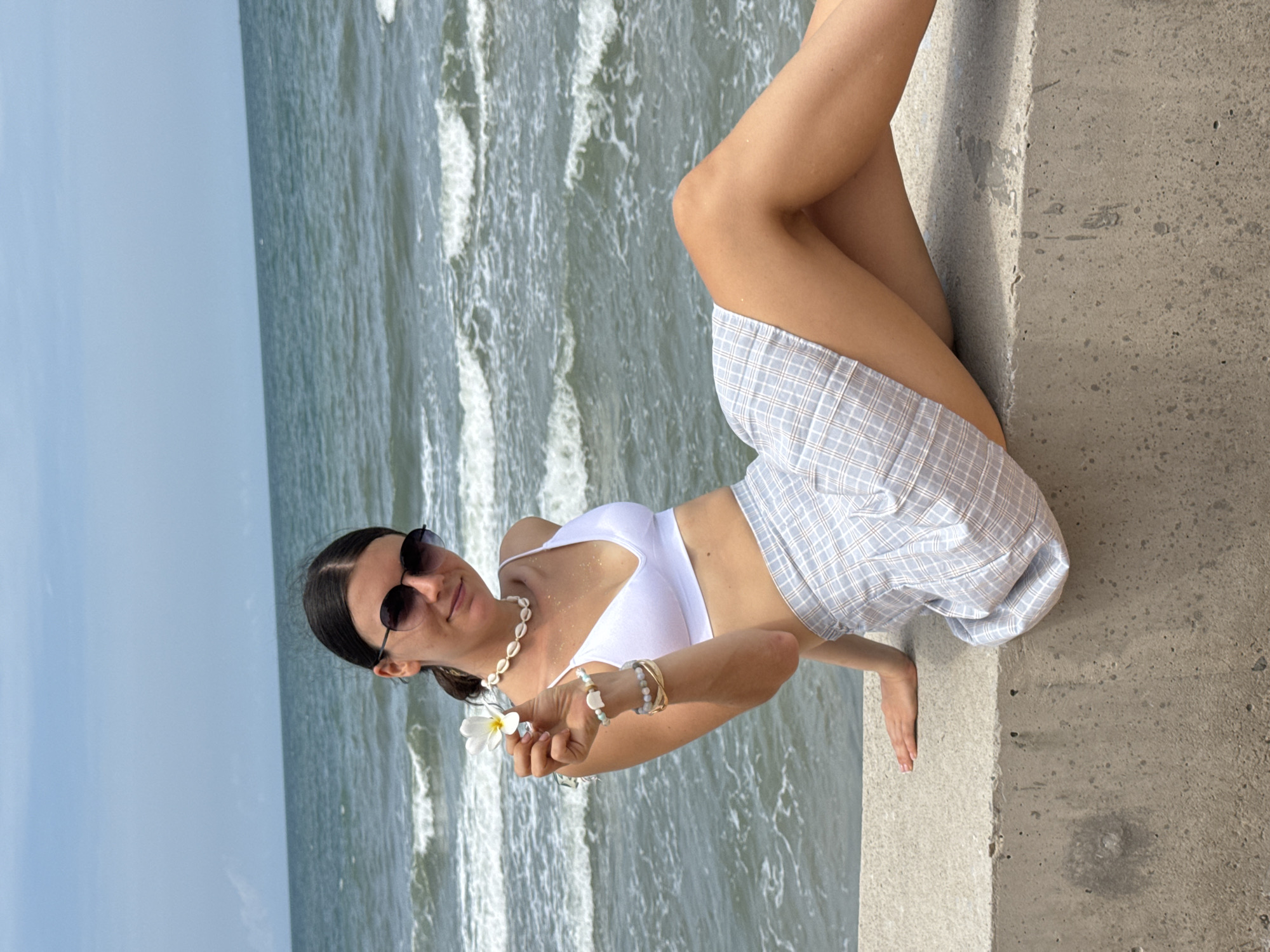 A young woman with dark hair pulled back and wearing sunglasses sits on a concrete ledge overlooking the ocean. She is dressed in a white bikini top and a light blue and white plaid skirt. In her left hand, she holds a white flower. She is adorned with a seashell necklace and several bracelets on her left wrist. The sea is visible in the background with waves breaking on the shore under a cloudy sky. The scene appears to be a coastal area in Mũi Né, Vietnam.
