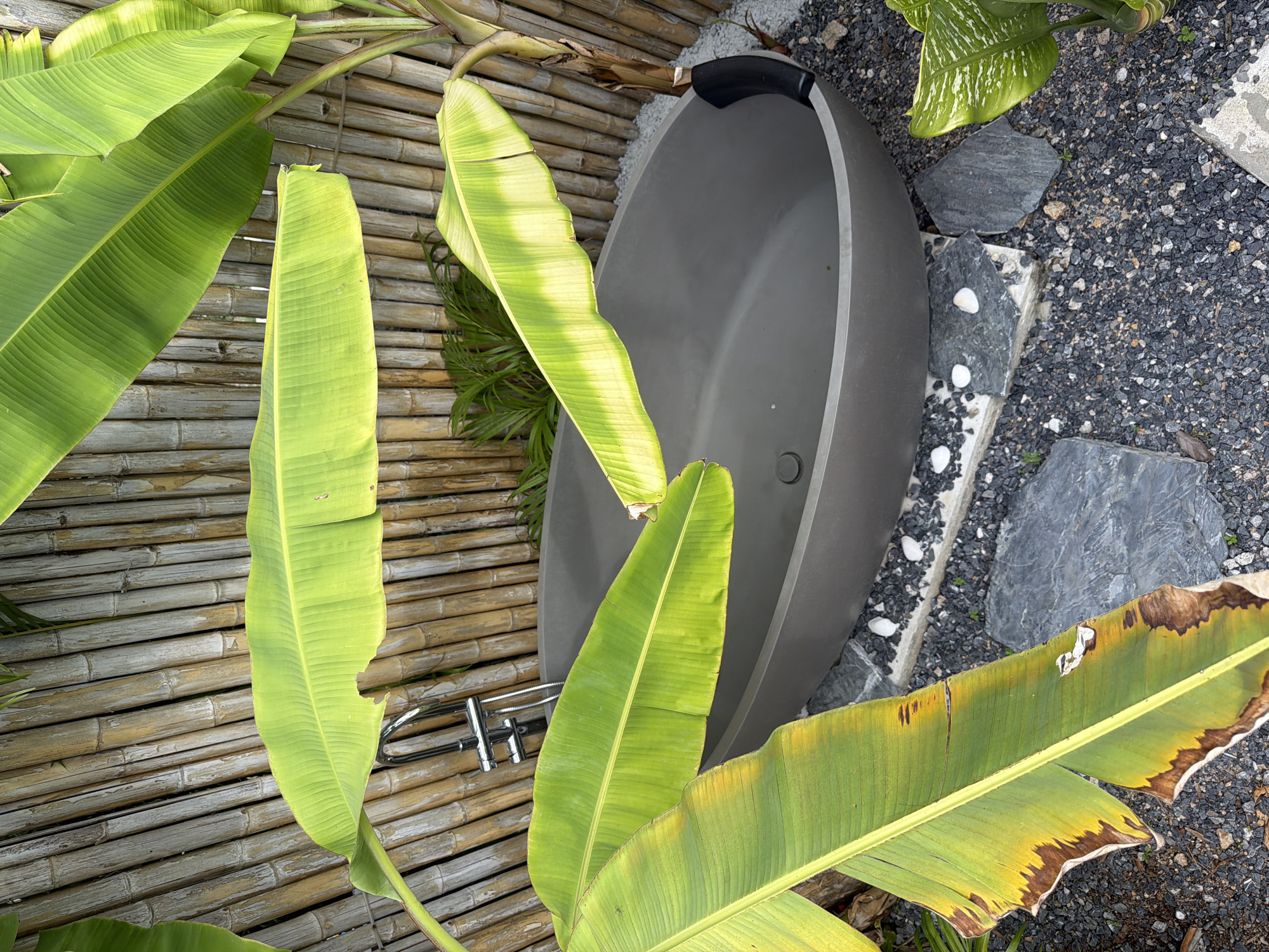 An outdoor bathroom area is shown in Baan Plai Laem, Thailand, featuring a grey, oval bathtub surrounded by dark gravel and flat stones. A bamboo fence serves as the backdrop, with large, green banana leaves arching over the scene. A chrome faucet and showerhead are visible behind the leaves, connected to a flexible hose.