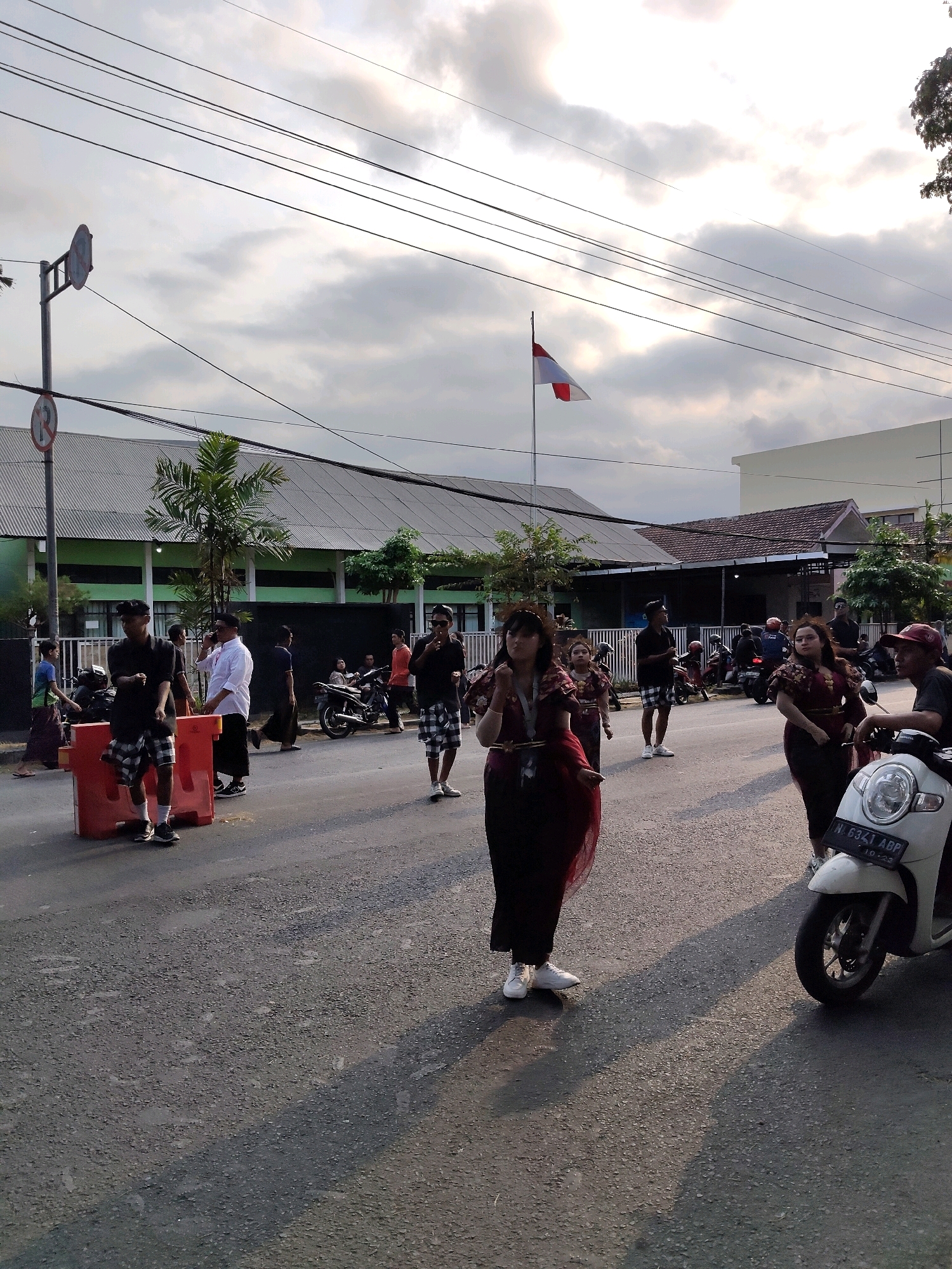 The image depicts a street scene in Malang, Indonesia, likely during a cultural event or parade given the traditional attire worn by some individuals. In the foreground, a woman in a dark red, flowing traditional dress with gold accents stands on the asphalt road. She is wearing white sneakers. Behind her, several other people, some also in traditional attire, are visible. A white scooter with a license plate is parked on the right side of the road. In the middle ground, a group of men are gathered, some wearing black shirts and checkered shorts, others in white shirts and dark pants. One man in a white shirt is on the phone. A red traffic barrier is placed on the left side of the road. In the background, buildings with green and grey facades line the street, and palm trees are visible. Overhead, power lines stretch across the cloudy sky, and an Indonesian flag is flying on a pole. The lighting suggests it is late afternoon, with long shadows cast across the road.