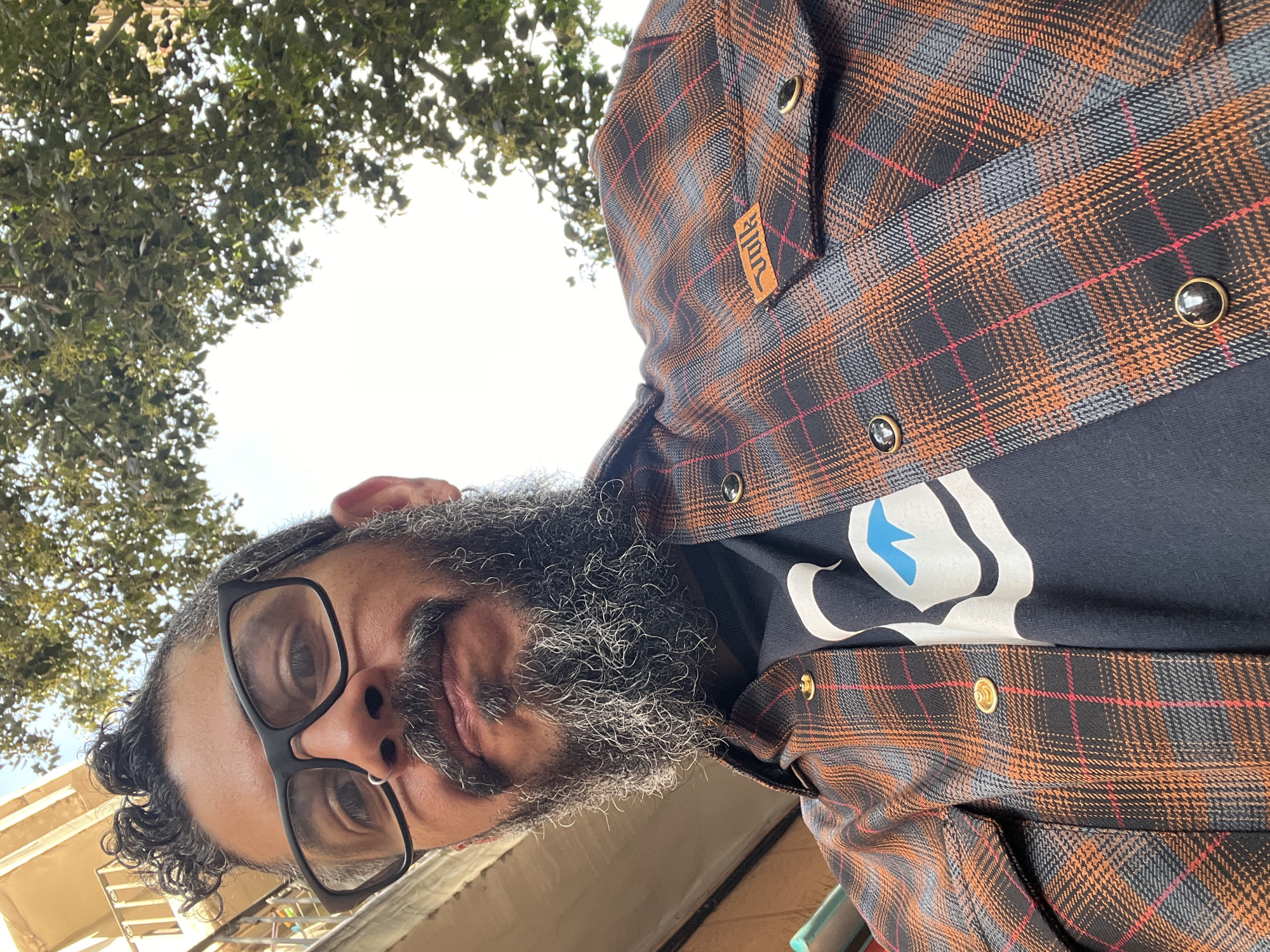 A man with graying hair and beard, wearing glasses and a plaid shirt over a black t-shirt with a white and blue design, is looking directly at the camera from a low angle. Trees are visible in the background, along with a partially visible building. The scene is in an urban environment. Without further information, the location in Barcelona cannot be verified.