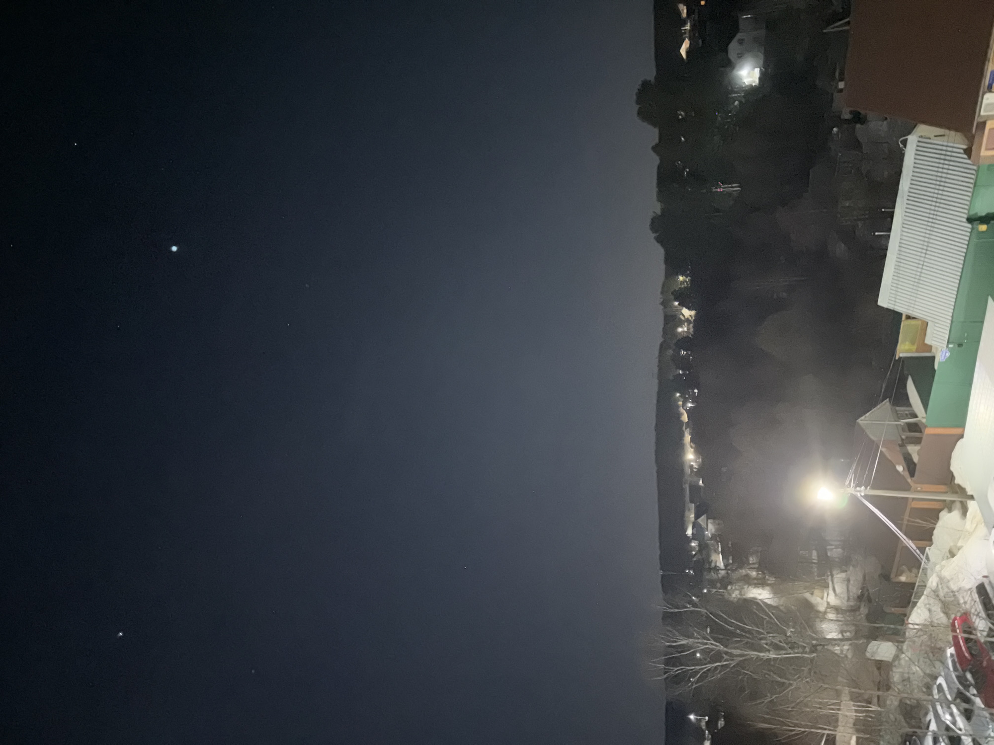 The image shows a nighttime scene in what appears to be Chaikovsky, Russia. The left side of the image is dominated by a dark sky, possibly a view over a body of water, with a few visible stars or distant lights. The right side of the image shows an urban or suburban area with buildings, trees, and streetlights. A bright streetlight illuminates a portion of the foreground, including bare tree branches and parked cars. The presence of the streetlight indicates an area with human activity. Other structures, likely residential or commercial buildings, can be seen in the background, illuminated by artificial lights. The overall impression is a clear night in a populated area.