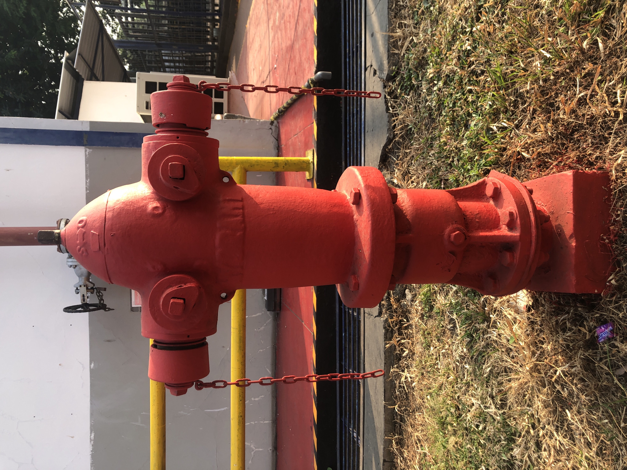 In Wonojati, Indonesia, a bright red fire hydrant stands prominently in the foreground, positioned on a patch of dry grass and sparse vegetation. The hydrant is a standard industrial design, featuring multiple outlets and caps, with red chains attached to the side caps hanging down. Behind the hydrant, a red paved surface with yellow and black striped markings is visible, suggesting a safety zone or walkway. A grey building with a white upper section forms the background, and a blue and white striped pillar is also present. The scene is outdoors, with sunlight casting shadows and illuminating the dry grass. A small piece of purple and silver candy wrapper is visible on the ground near the base of the hydrant. Beyond the building, a metal fence and trees can be seen, indicating an urban or industrial setting.