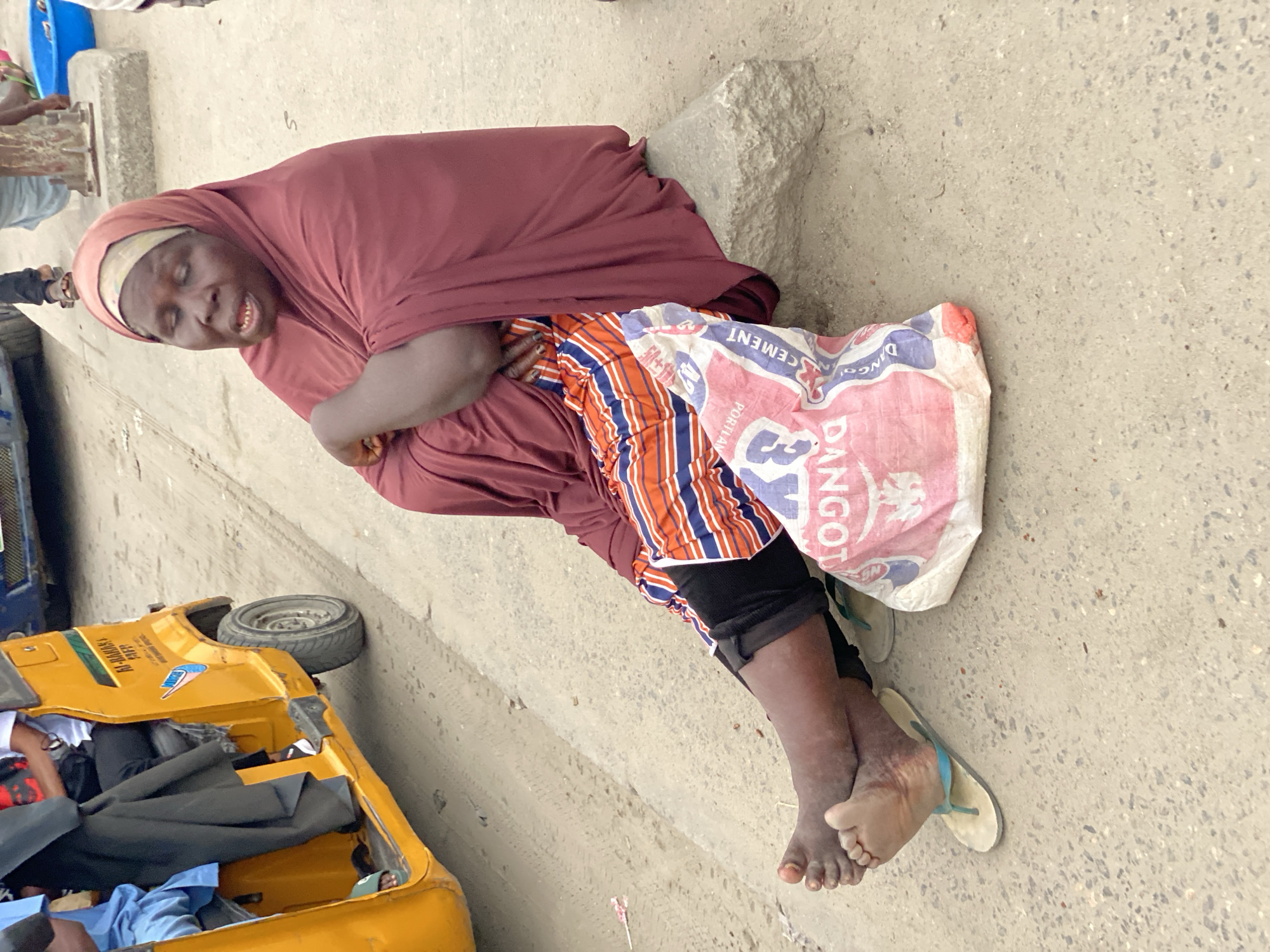 A woman, likely an elderly adult, is lying on the ground next to a yellow tricycle (Keke Napep), a common mode of transportation in Lagos, Nigeria. The woman is wearing a maroon hijab and an orange, white, and blue patterned skirt. She is lying on a bag of Dangote Cement. She has bare feet and is wearing flip-flops. The ground is concrete. The location is likely in Lagos, Nigeria, given the presence of the Keke Napep and the Dangote Cement bag.