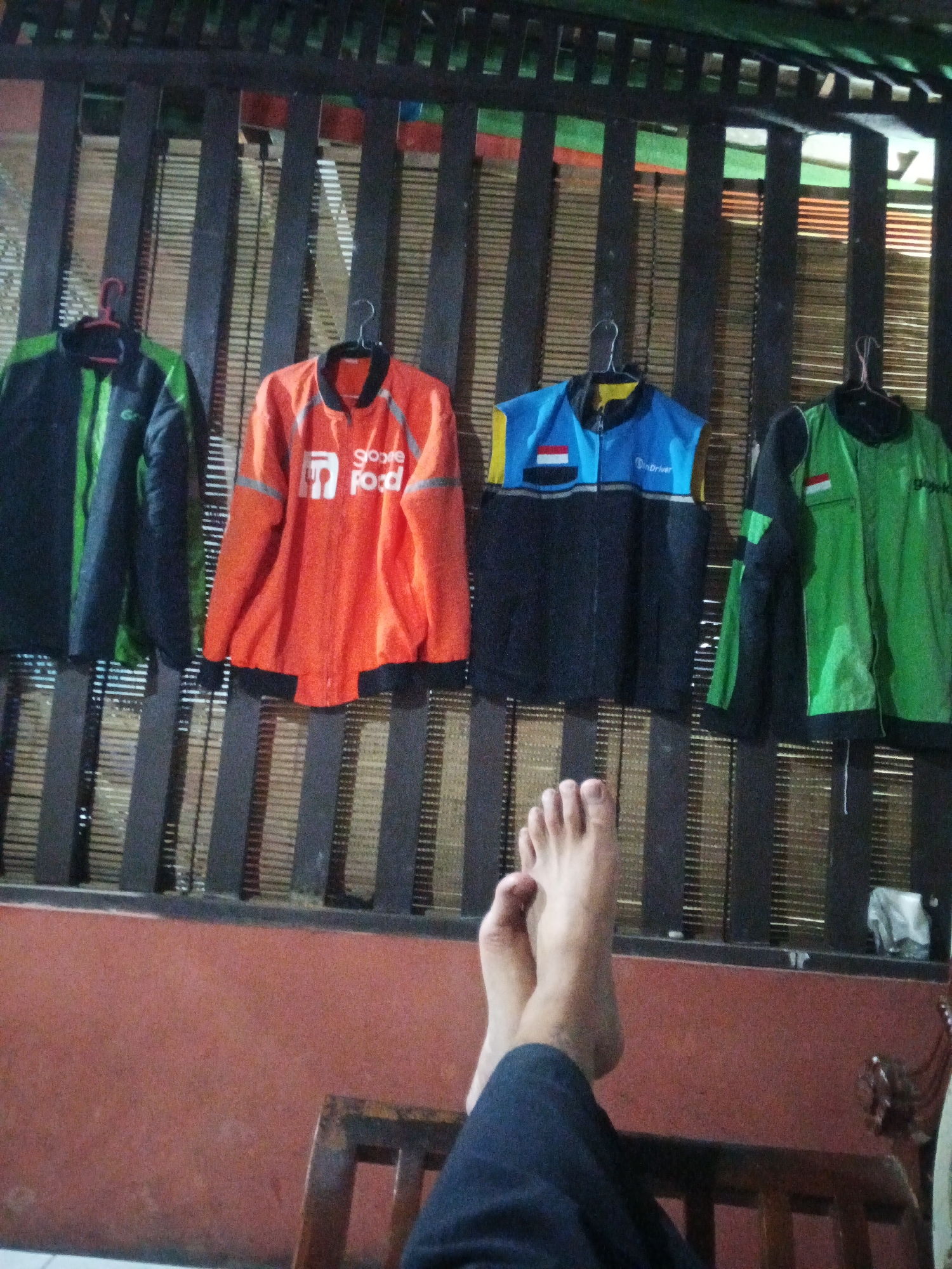 The image shows four jackets and vests hanging on a wooden slatted wall. The clothing includes a dark green and black jacket, an orange jacket with "Google Food" branding, a blue and black vest with "GoDriver" branding and an Indonesian flag motif, and a green and black jacket. In the foreground, a person's legs and bare feet are crossed, resting on a wooden chair or furniture. The background is a bamboo-like textured wall. The scene appears to be indoors, possibly a storage area or a casual living space. The clothing suggests a connection to delivery services, possibly in Purwanegara, Indonesia, given the Indonesian flag motif.