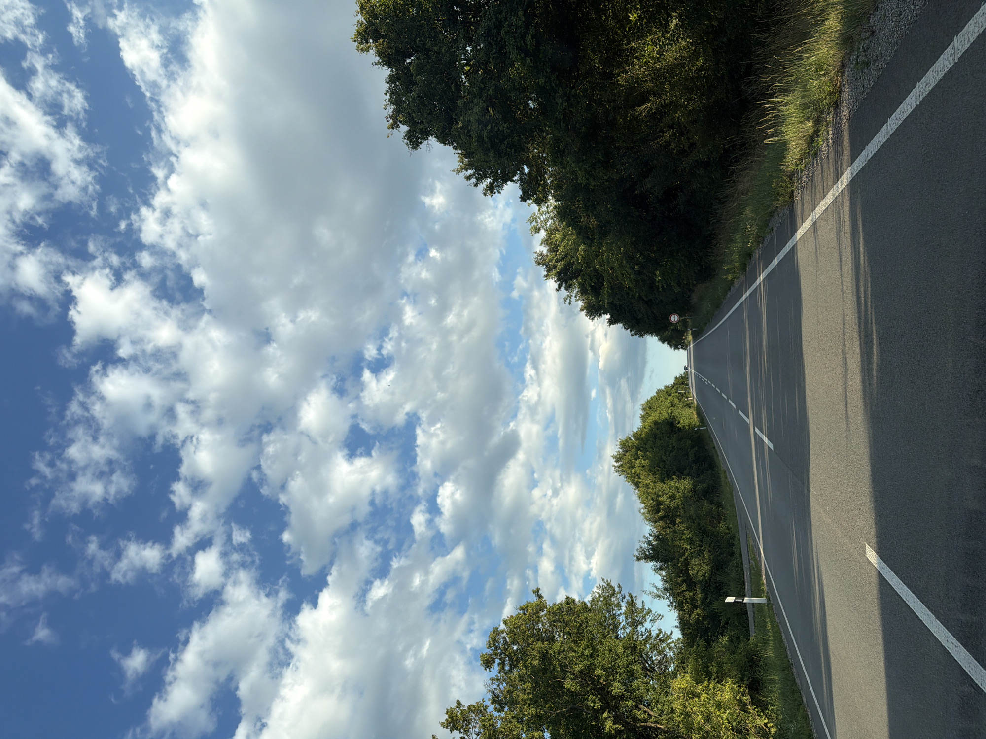 The image shows a rural asphalt road in the municipality of Veľaty, Slovakia, on a clear day with scattered white clouds against a blue sky. The road stretches into the distance, flanked by lush green trees and vegetation on both sides. White lines mark the edges and center of the road. A circular red and white traffic sign is visible on the right side of the road in the distance. The sunlight casts shadows across the asphalt, indicating the time of day is likely afternoon. There are no people or vehicles visible in the image.