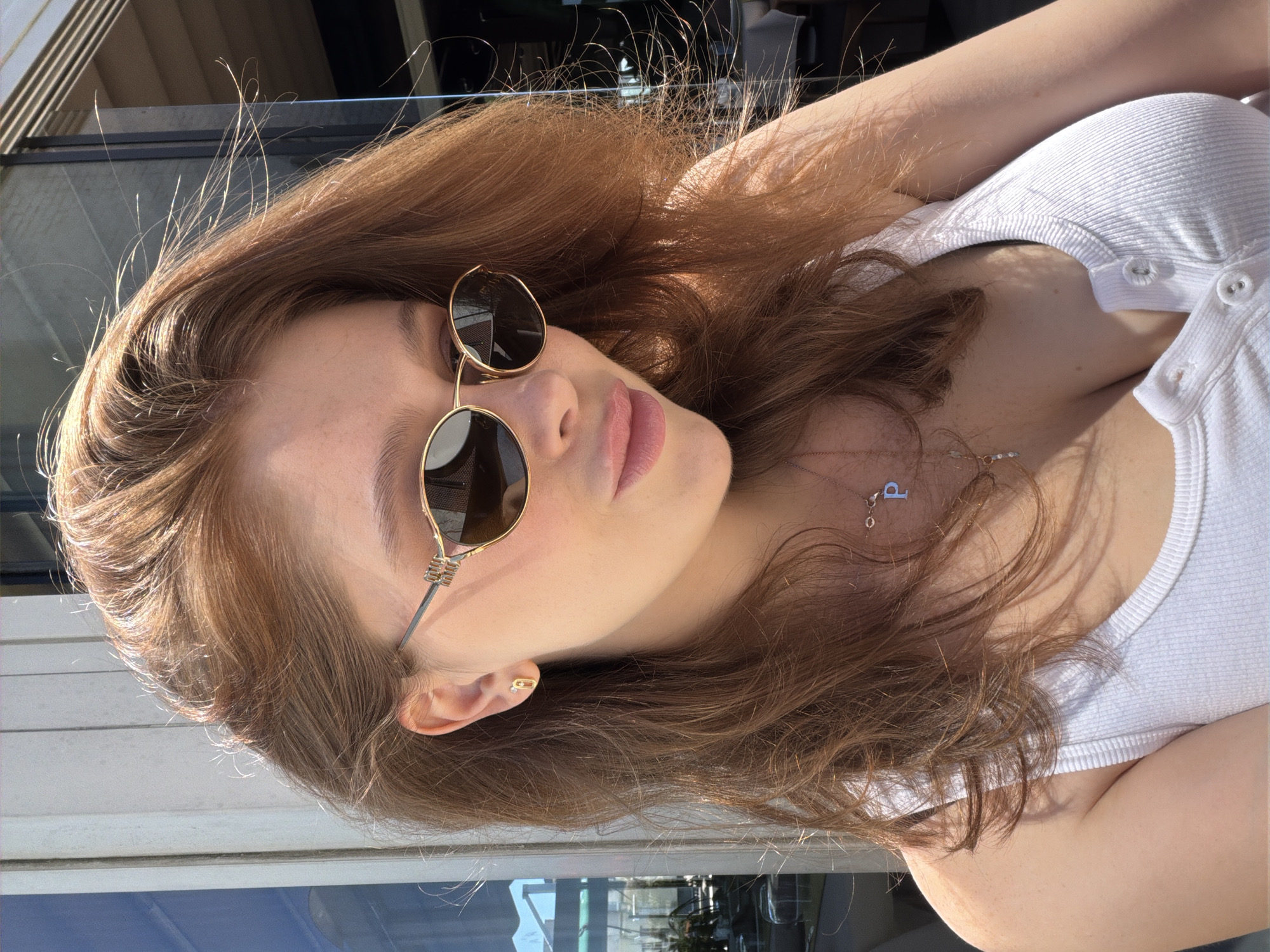 A young woman with brown hair and fair skin is the central subject of the image, taken from an elevated perspective. She is wearing a white ribbed tank top and round sunglasses with a gold frame. A gold necklace with a pendant is visible around her neck, and she has a gold earring. The background suggests an urban setting. The presence of reflections and materials suggests an outdoor seating area of a cafe or restaurant, with modern architectural elements. It is not possible to determine from this image if it is in Istanbul or anywhere else, and Istanbul is not discernible from this photo.