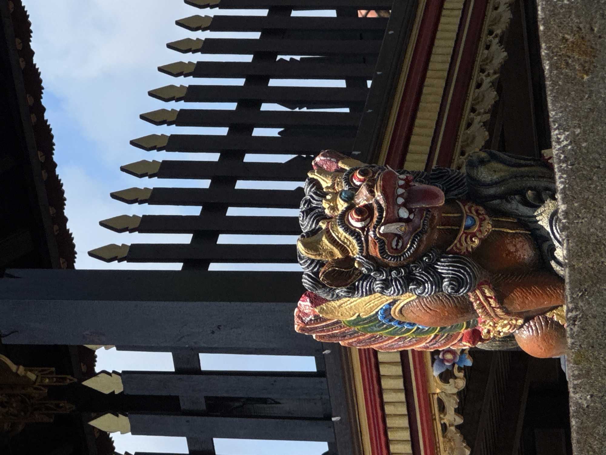 The image presents a close-up, upward-angled view of an ornate, multi-colored carving integrated into the architecture of a traditional building, likely in North Kuta, Indonesia. The central subject is a vibrant, mythical guardian figure, possibly a Barong or a protective lion, depicted with an open mouth revealing teeth, prominent eyes, and an elaborate mane or frills. The carving is richly detailed with hues of red, gold, black, and blue, suggesting it serves as a decorative corbel or support.
Above and to the left of the carving, a series of dark wooden beams or slats are arranged in a tiered pattern, forming part of a roof or ceiling structure. Many of these slats are adorned with pointed, golden tips. Through the gaps in this wooden framework, a bright sky with scattered clouds is visible, indicating that the photo was taken during the day. To the far right, a textured, rough-hewn stone pillar or column is partially visible, alongside other gilded and red-striped architectural elements. The intricate craftsmanship and style of the carving and surrounding structure are highly characteristic of Balinese art and traditional architecture. No specific activity is taking place, and no text is visible in the frame.