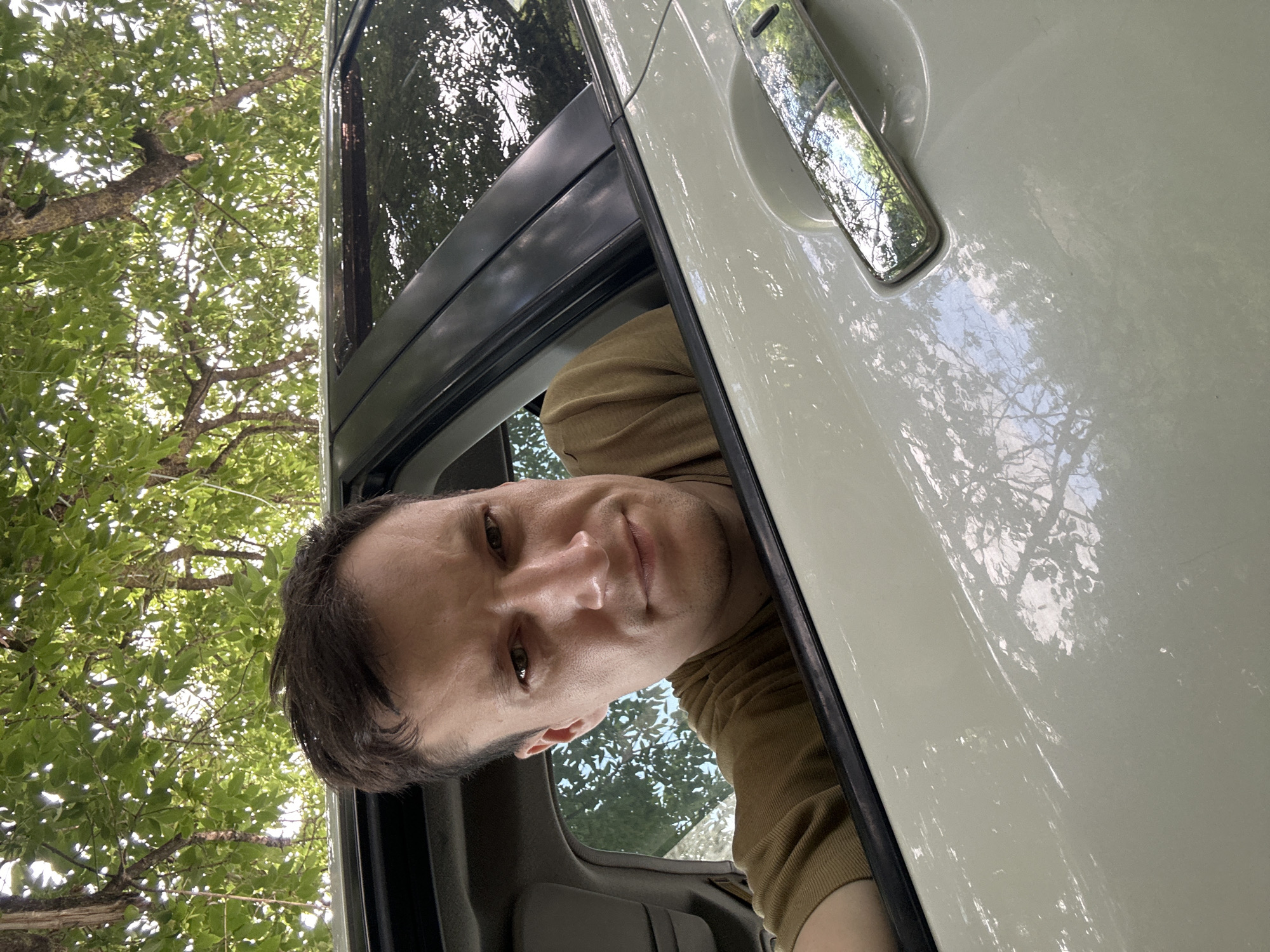 A man is looking directly at the camera from inside a car. He is wearing a tan shirt and has short brown hair. The car is light-colored and appears to be parked under trees, as evidenced by the foliage and dappled sunlight reflecting on the car's surface. The photo appears to have been taken outdoors during the daytime. The specific location in Makiv, Ukraine, is not discernible from the image itself.