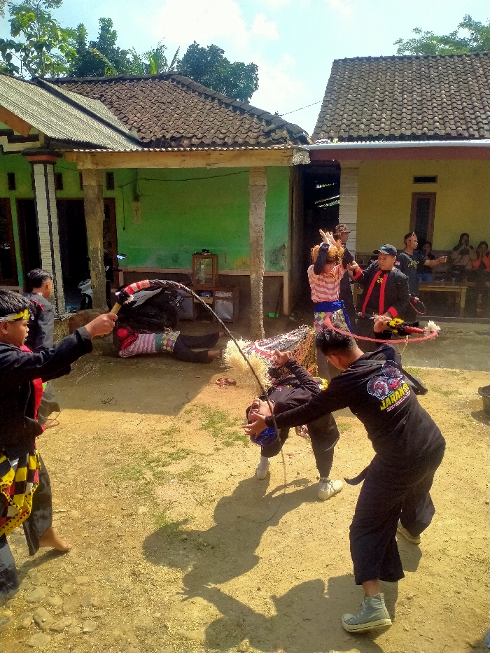 The image depicts an outdoor cultural performance in Glonggong, Indonesia, set against the backdrop of two single-story houses under a clear sky. The ground is dry, light brown earth with sparse green patches.
In the foreground, four individuals are actively involved in the performance. To the left, a male in a black shirt with red trim, dark pants, and a yellow and black patterned sarong is barefoot, holding a whip (pecut) extended forward. Centered, a person lies prone on the ground, wearing a pink and white striped shirt and dark pants, partially covered by a dark fabric. To their right, another performer, dressed in a black long-sleeved shirt and dark pants, is bent dramatically backward, head near the ground, with a blue object covering their face and mouth open. A whip's lash appears to encircle their neck/head. Directly behind this person, a male in a black long-sleeved shirt with "JARANAN" printed on the back, dark pants, and light-colored shoes, holds a whip with its lash extending towards the bent-over performer.
In the mid-ground, near the green-painted house to the left, a person is partially visible. Further back, near the right, yellow-painted house, a man in a black jacket with red trim and a black cap stands, gesturing. Adjacent to him, a woman in a striped shirt gestures with both hands raised. Several other individuals are seated on a wooden bench in the background, observing the scene.
The houses feature tiled roofs and light-colored walls (green on the left, yellow on the right). The left house has concrete pillars supporting a corrugated metal overhang, and a wooden cabinet with a glass top is visible on its porch. Shadows indicate bright, direct sunlight.