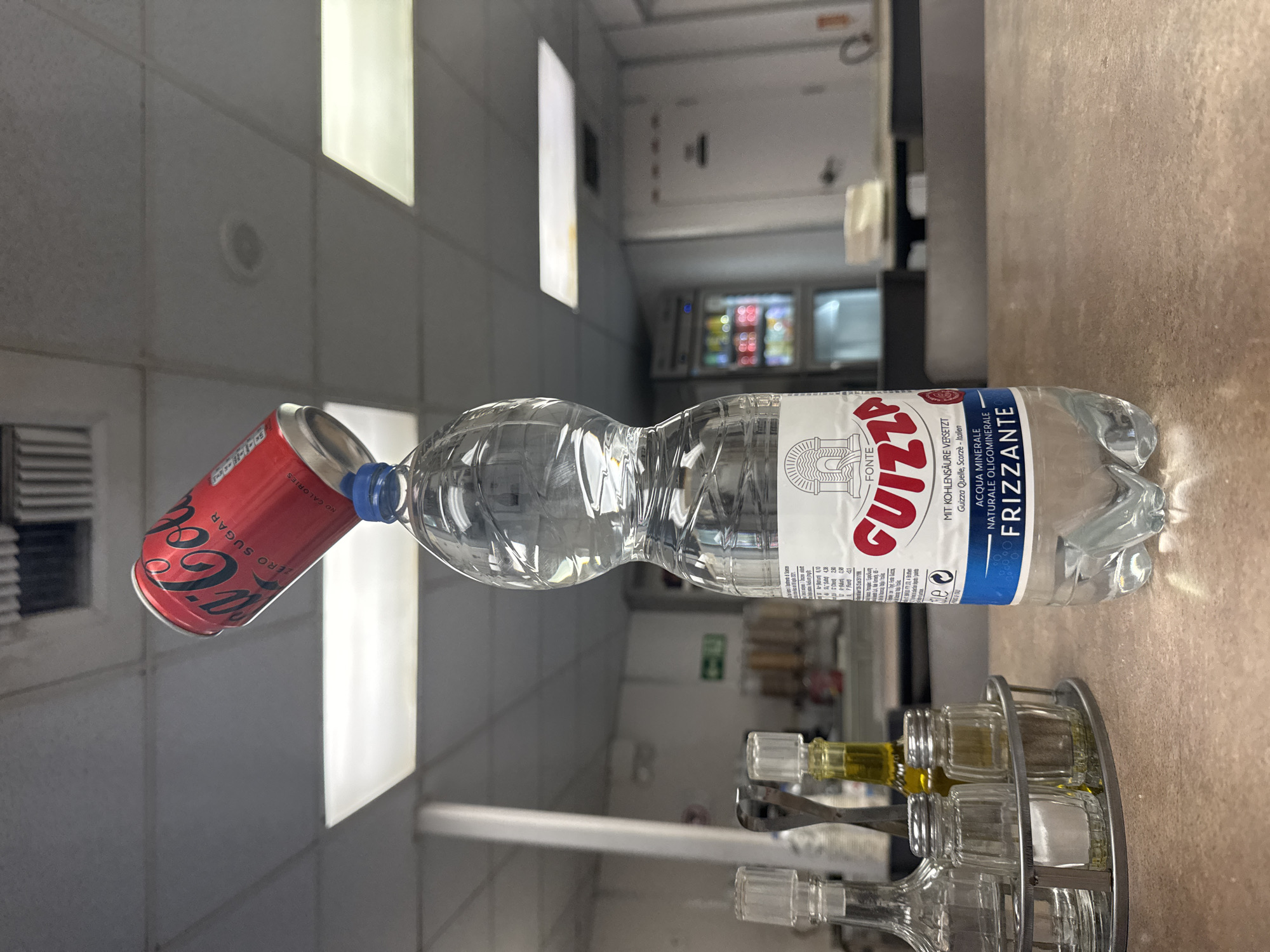 A red Coca-Cola Zero Sugar can is precariously balanced on the blue cap of a clear plastic bottle of Guizza Frizzante Acqua Minerale. The bottle is resting on a brown countertop. In the background, a refrigerated display case filled with colorful beverages is visible. The scene appears to be indoors, possibly in a cafe or convenience store, with fluorescent lighting and acoustic ceiling tiles. There are no people visible in the image.