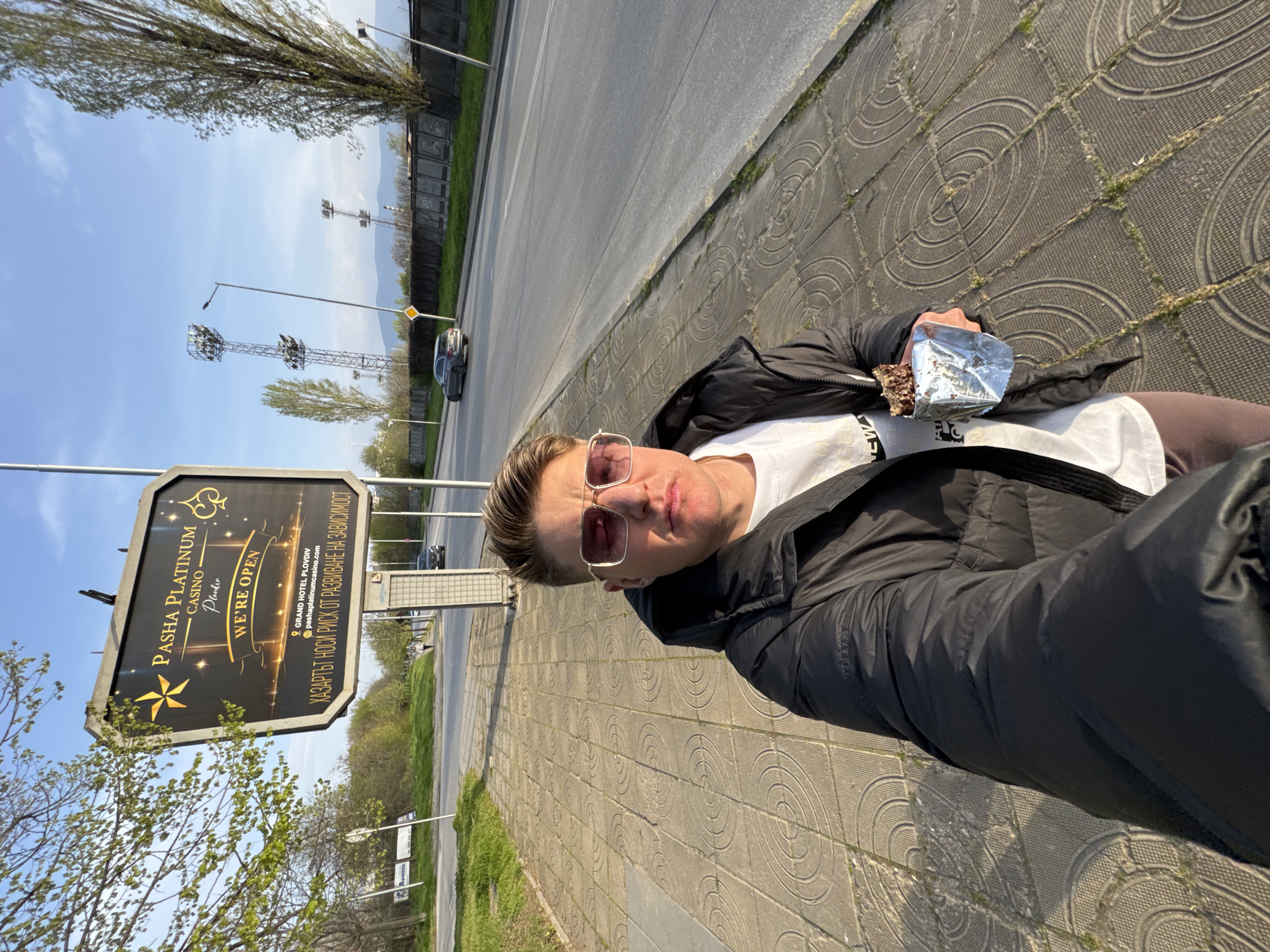 A selfie of a man standing on a sidewalk next to a road with a large sign for "Pasha Platinum Casino" in Plovdiv, Bulgaria. He is wearing a white t-shirt, a black jacket, and sunglasses, and he is holding a partially unwrapped chocolate bar in foil in his left hand. The background shows a multi-lane road with cars, trees, and utility poles. The sidewalk has a circular pattern. The sky is partly cloudy.